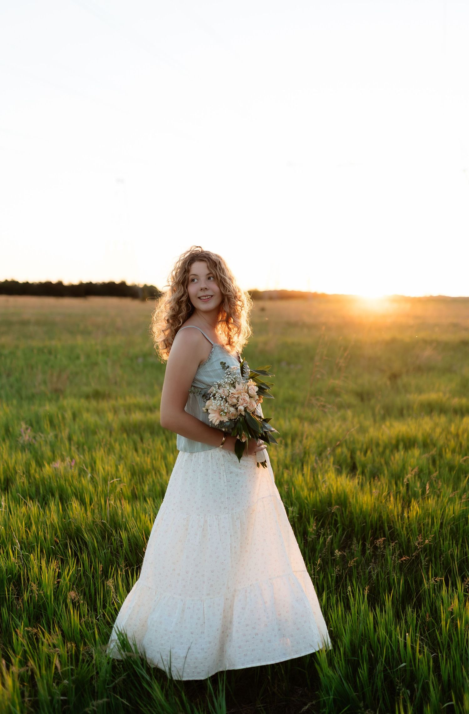 Curly-haired woman holding wildflower bouquet in white dress at golden sunset in field.