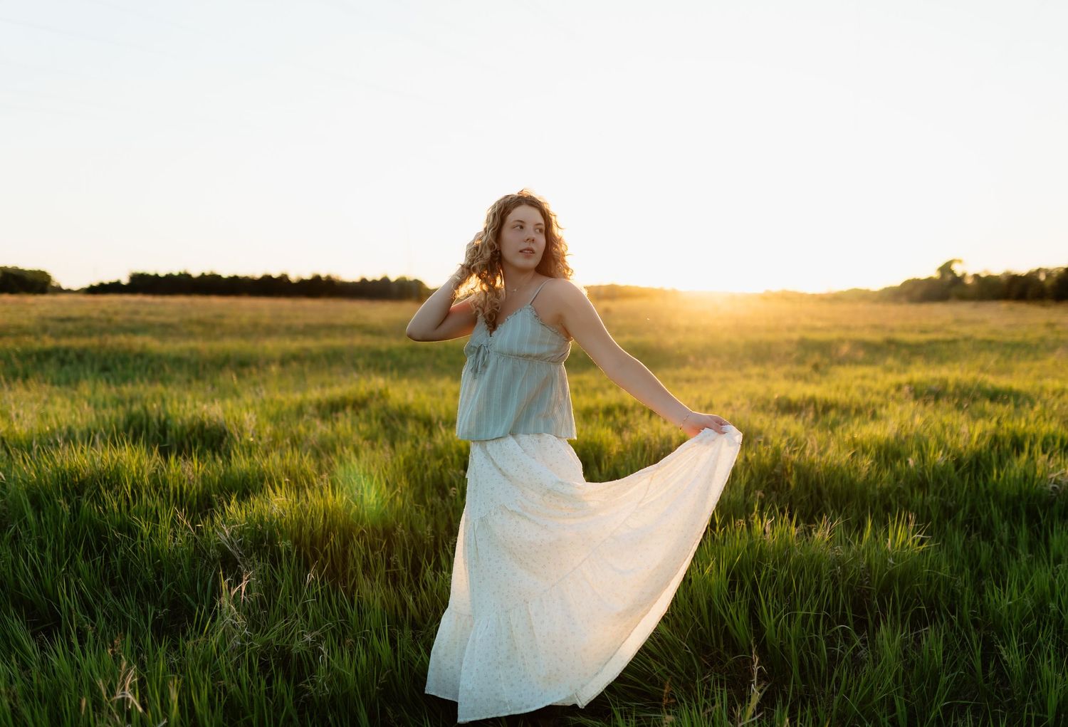 Young woman in light blue top and white skirt twirling in a green field during a vibrant sunset.