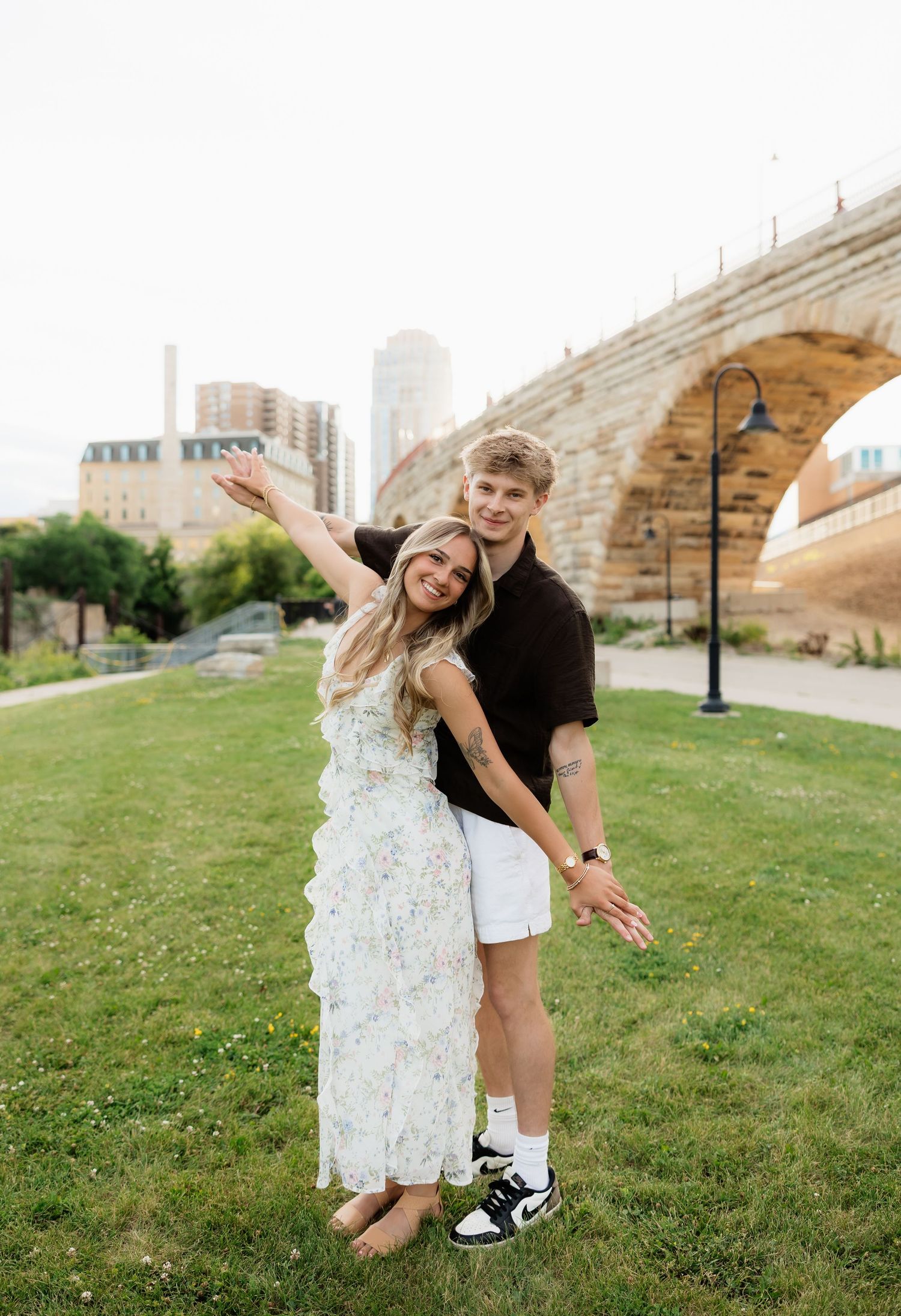 Joyful couple laughing and posing playfully on green lawn with historic stone arch bridge in the background.