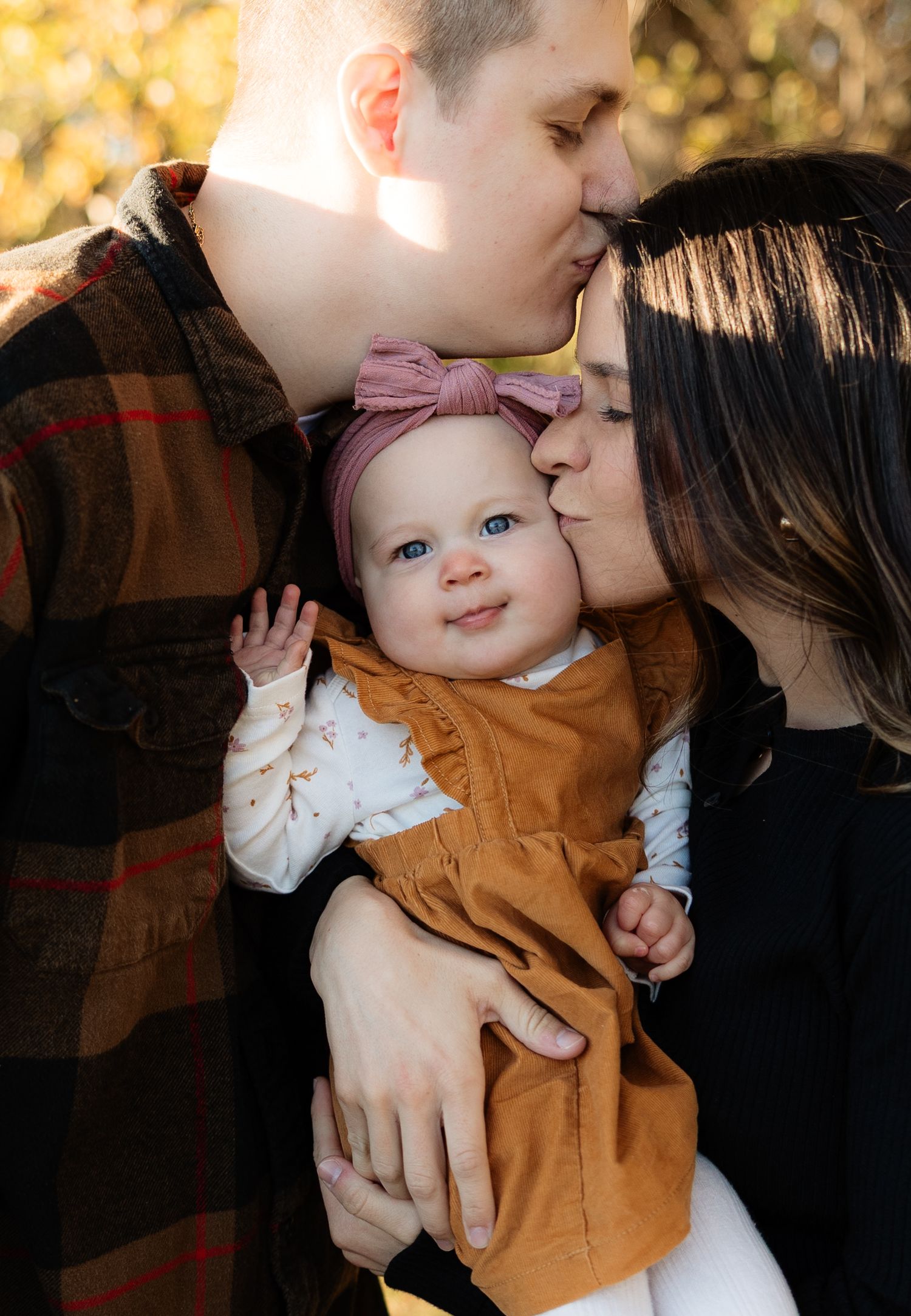 Parents kissing their smiling blue-eyed baby girl wearing a pink bow in autumn light.