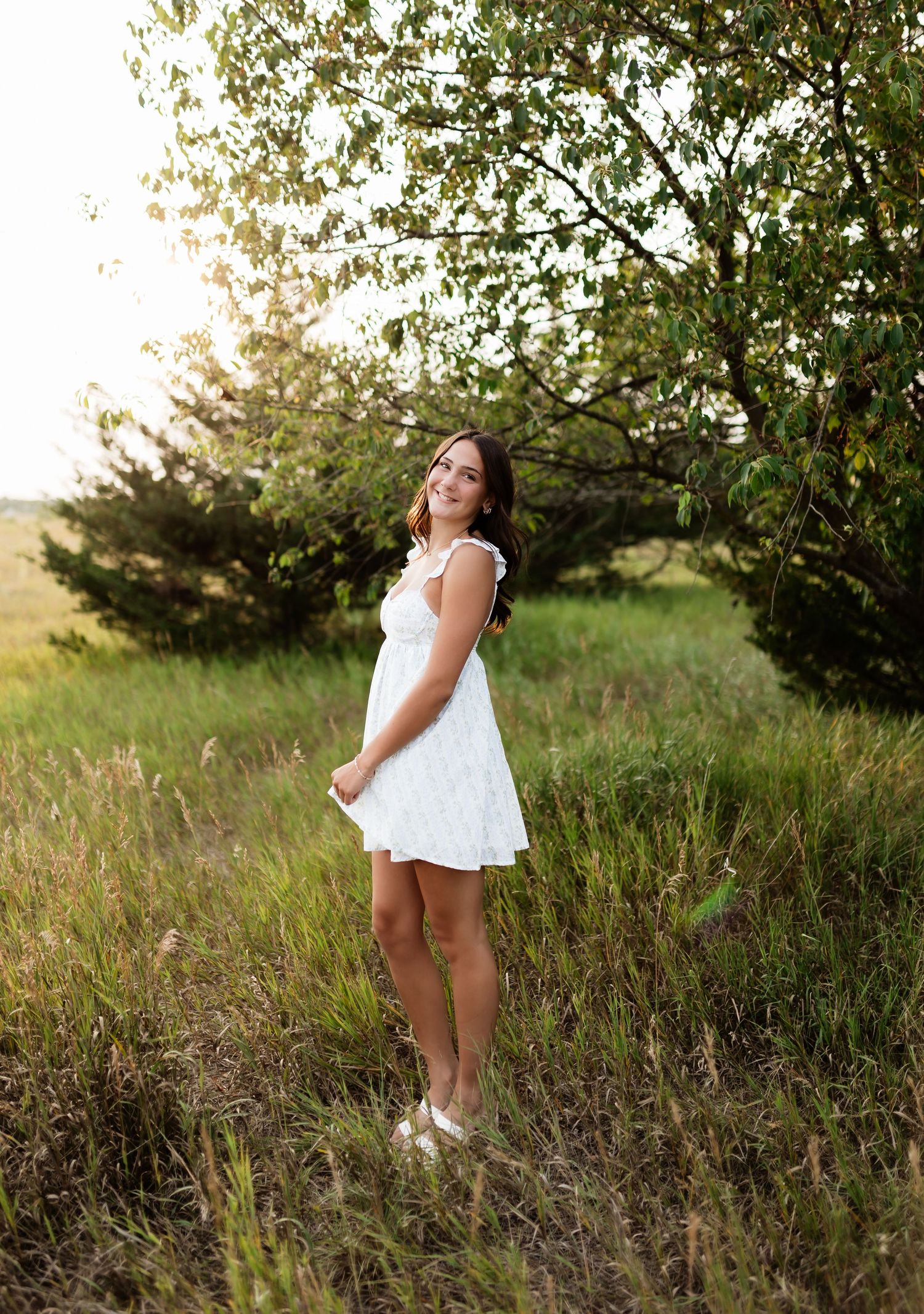 Young woman in white dress standing in golden meadow with sunlit trees in background.