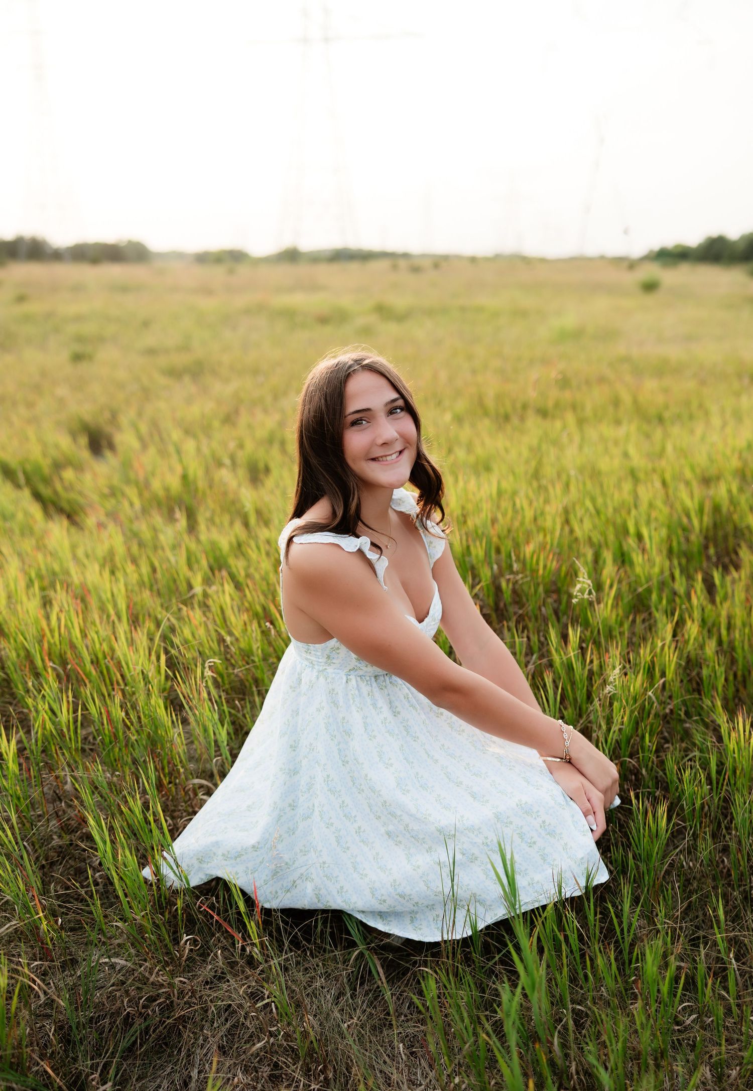 Smiling young woman in a light blue floral sundress crouching in a vast green field at golden hour.