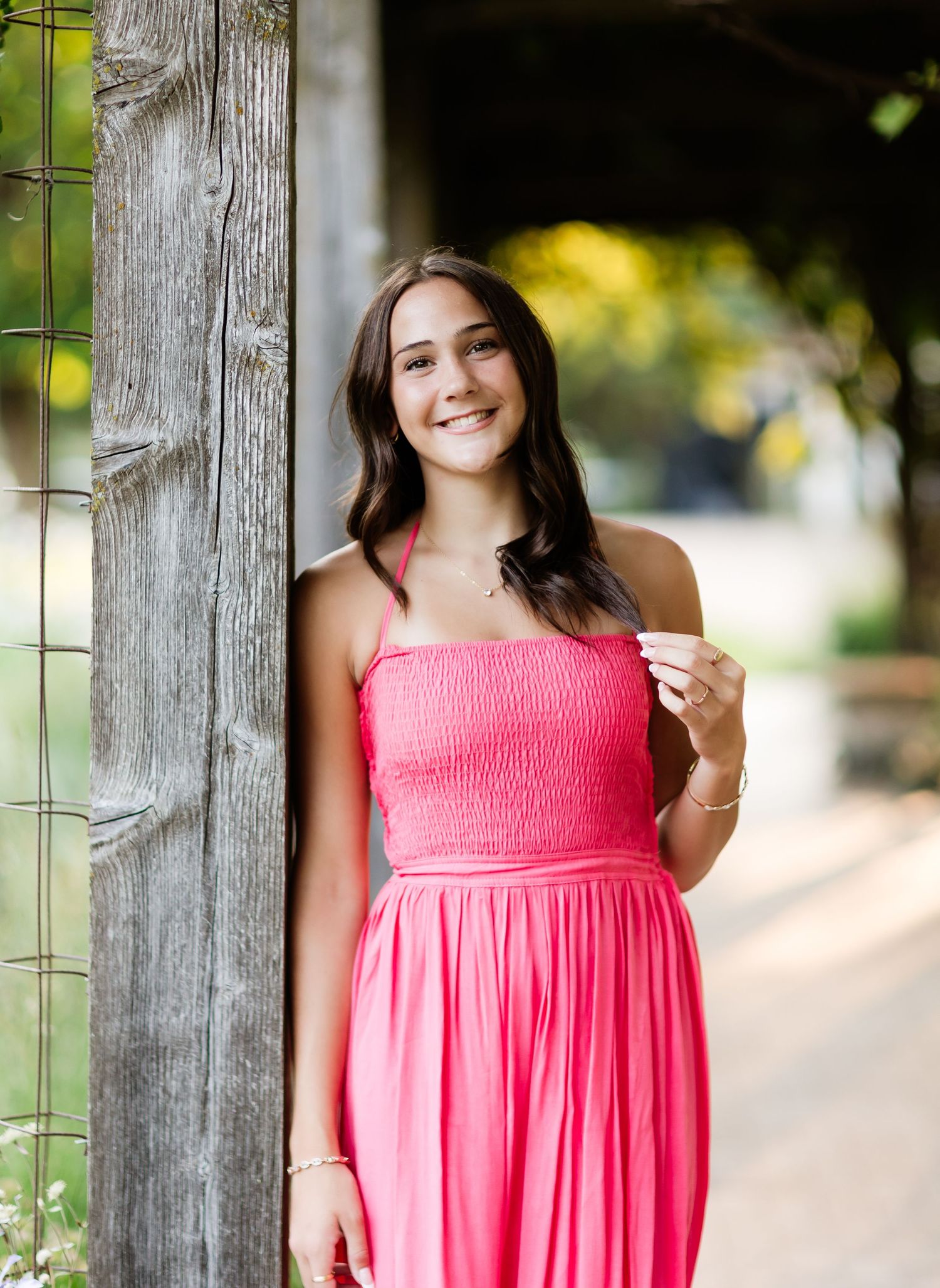Smiling young woman in pink maxi dress leaning against rustic wooden post outdoors.