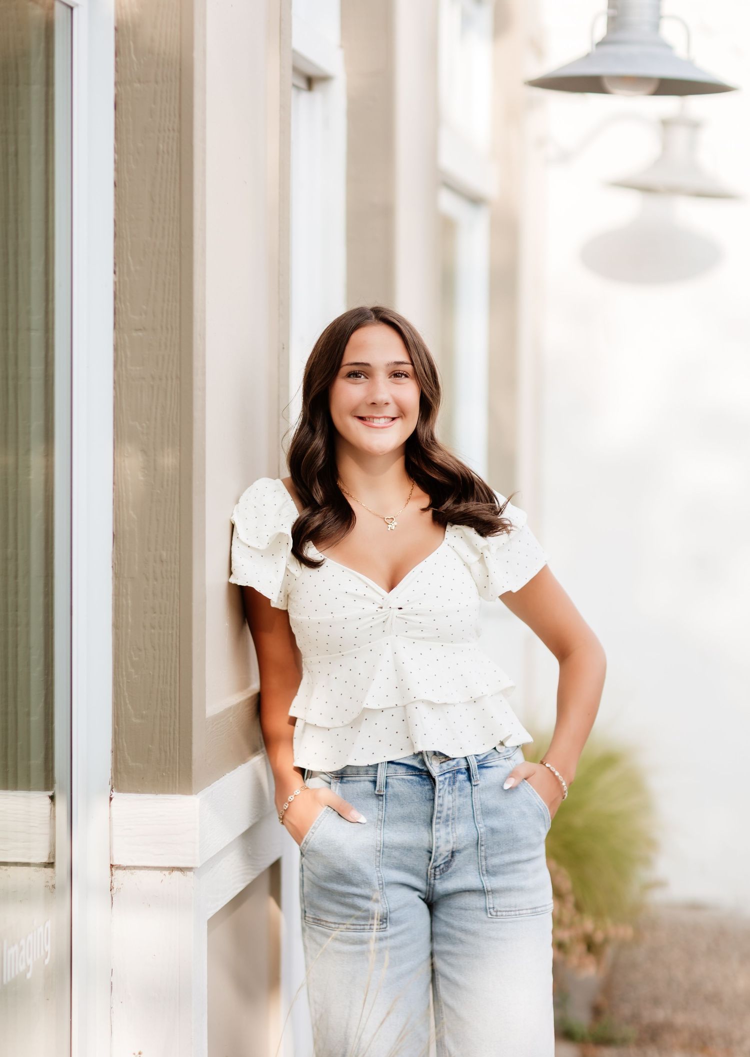 Smiling brunette woman in white ruffled crop top and jeans leaning against a building exterior.