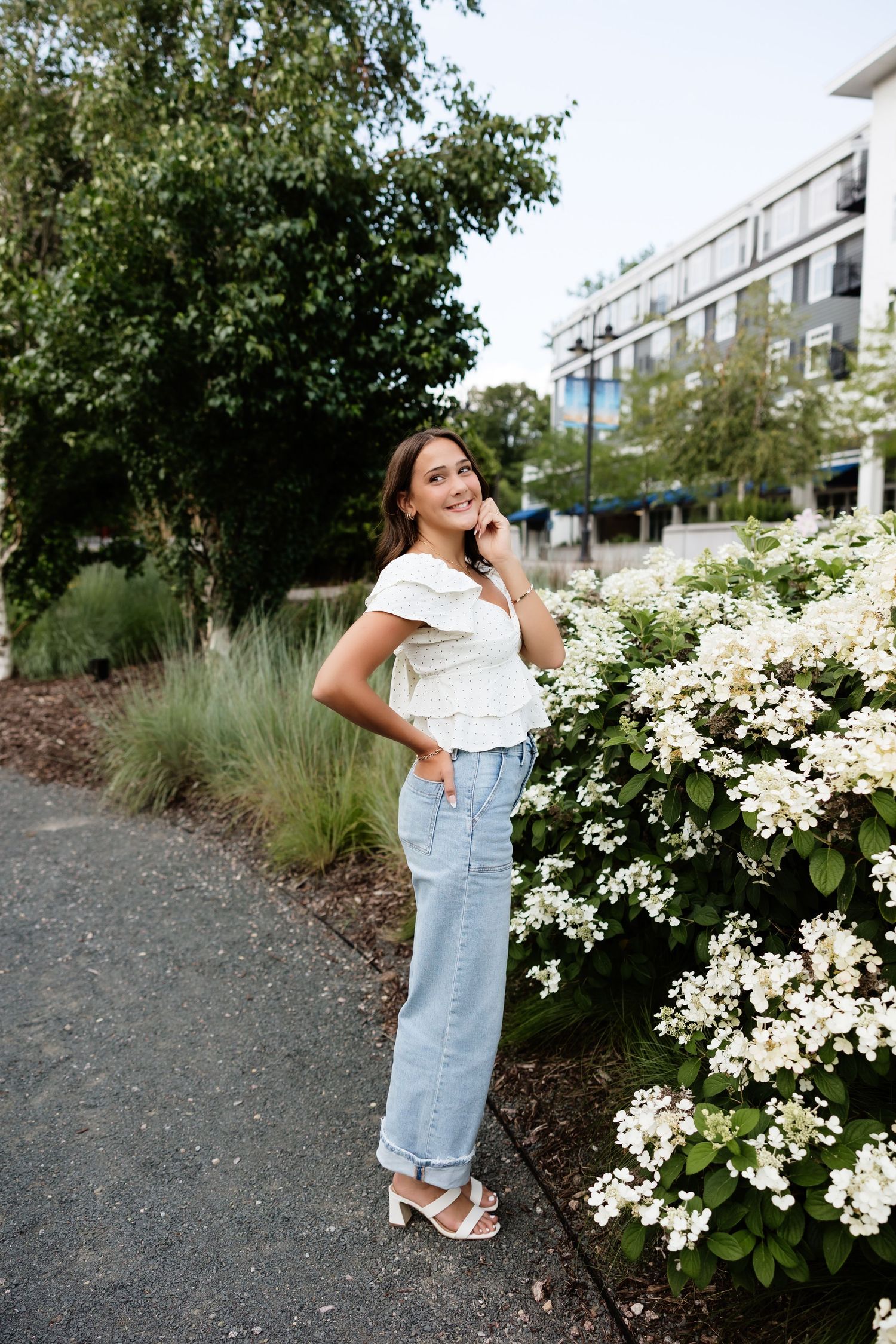 Young woman in white top and wide-leg jeans posing beside white hydrangea blooms in an urban garden setting.