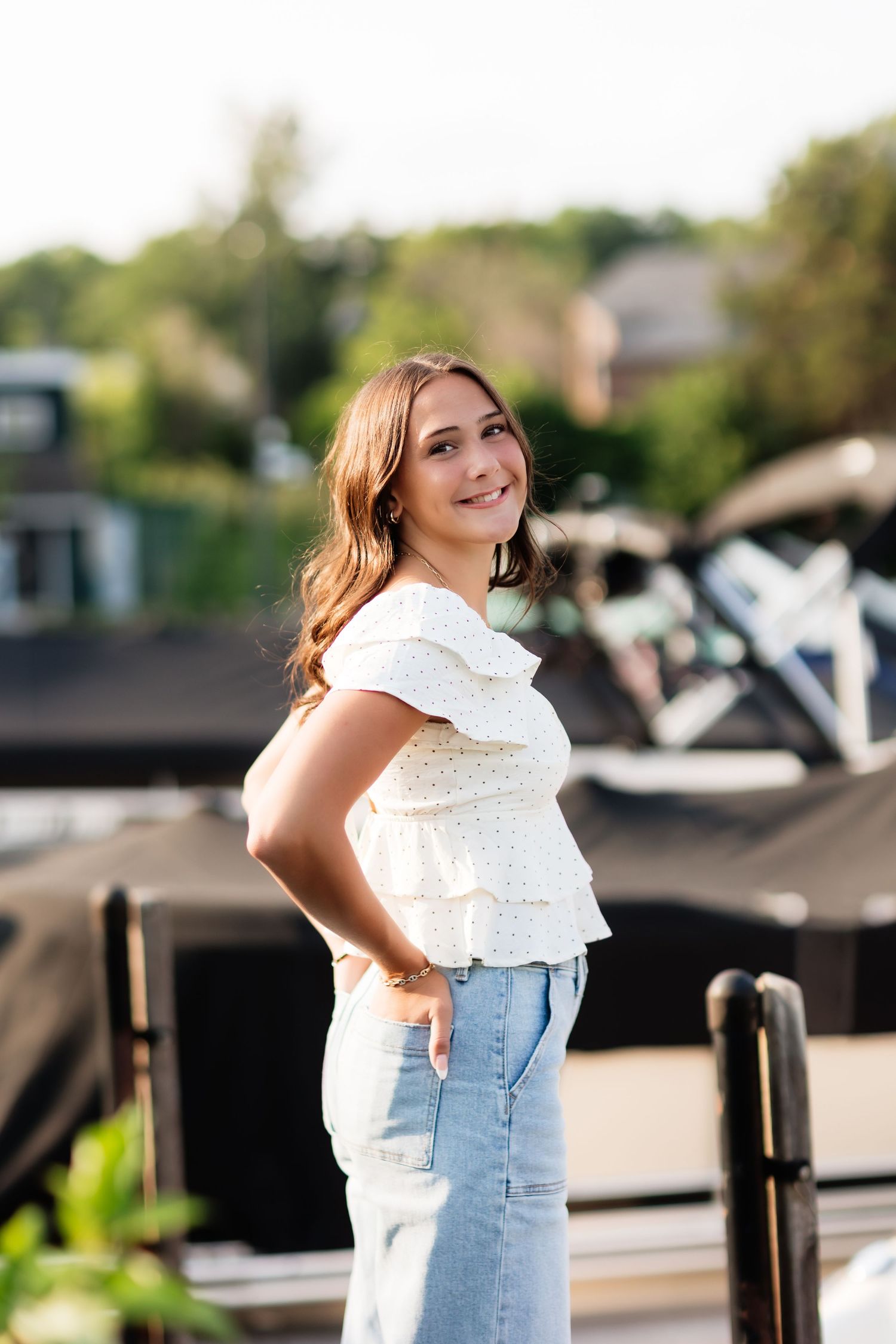 Smiling young woman in white ruffled top and light jeans posing outdoors near a marina dock.