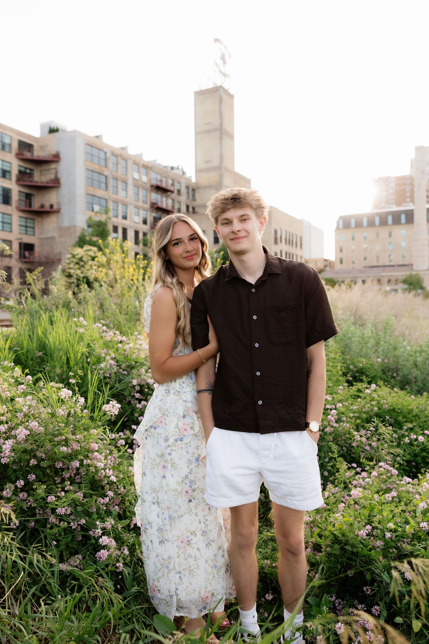 Smiling young couple posing together in a wildflower garden with urban buildings behind them.