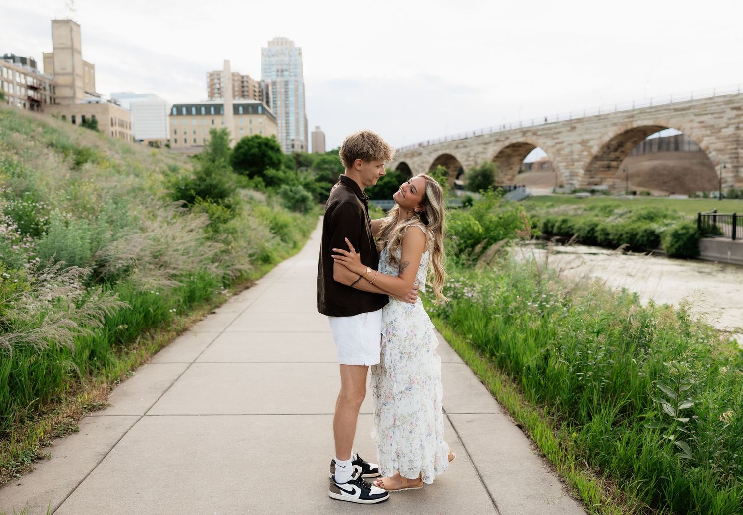 A couple sharing a romantic moment on a riverside path with lush greenery and a stone arch bridge in the background.