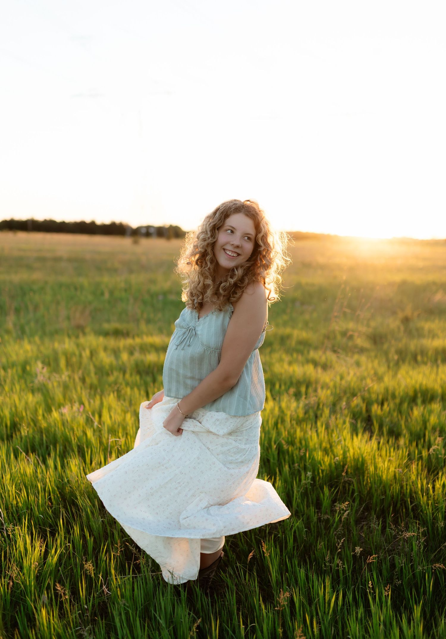 Joyful curly-haired woman twirling in a white dress in a golden sunset field.