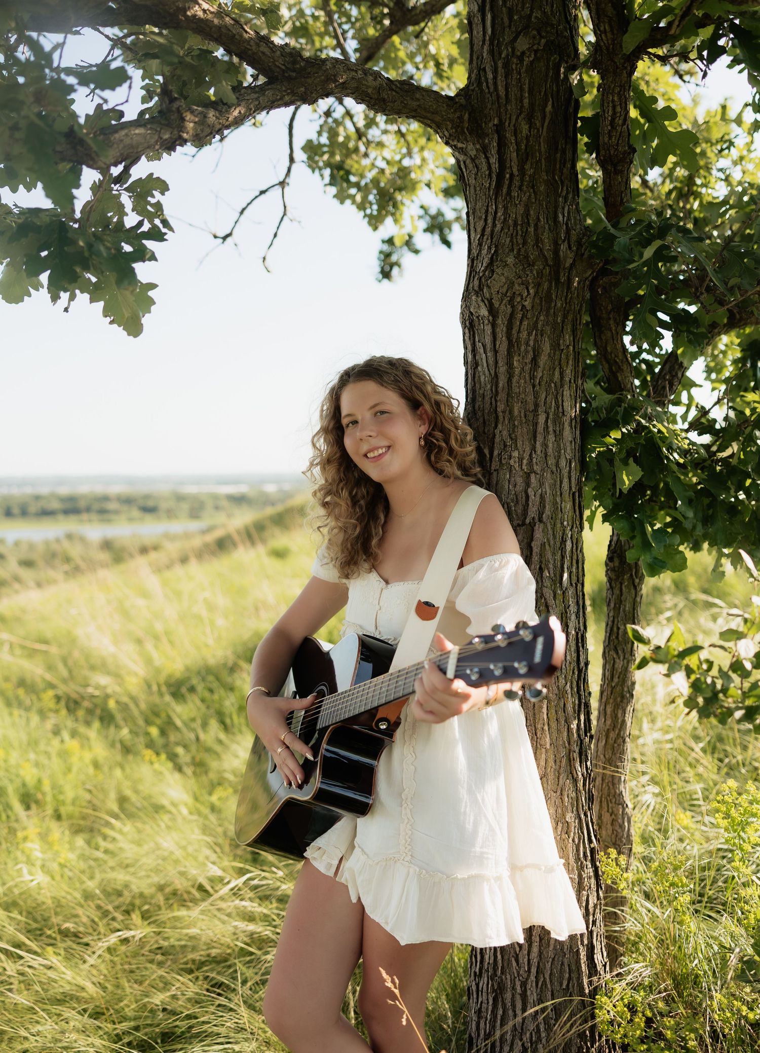 Smiling young woman in white dress playing acoustic guitar while leaning against a tree outdoors.