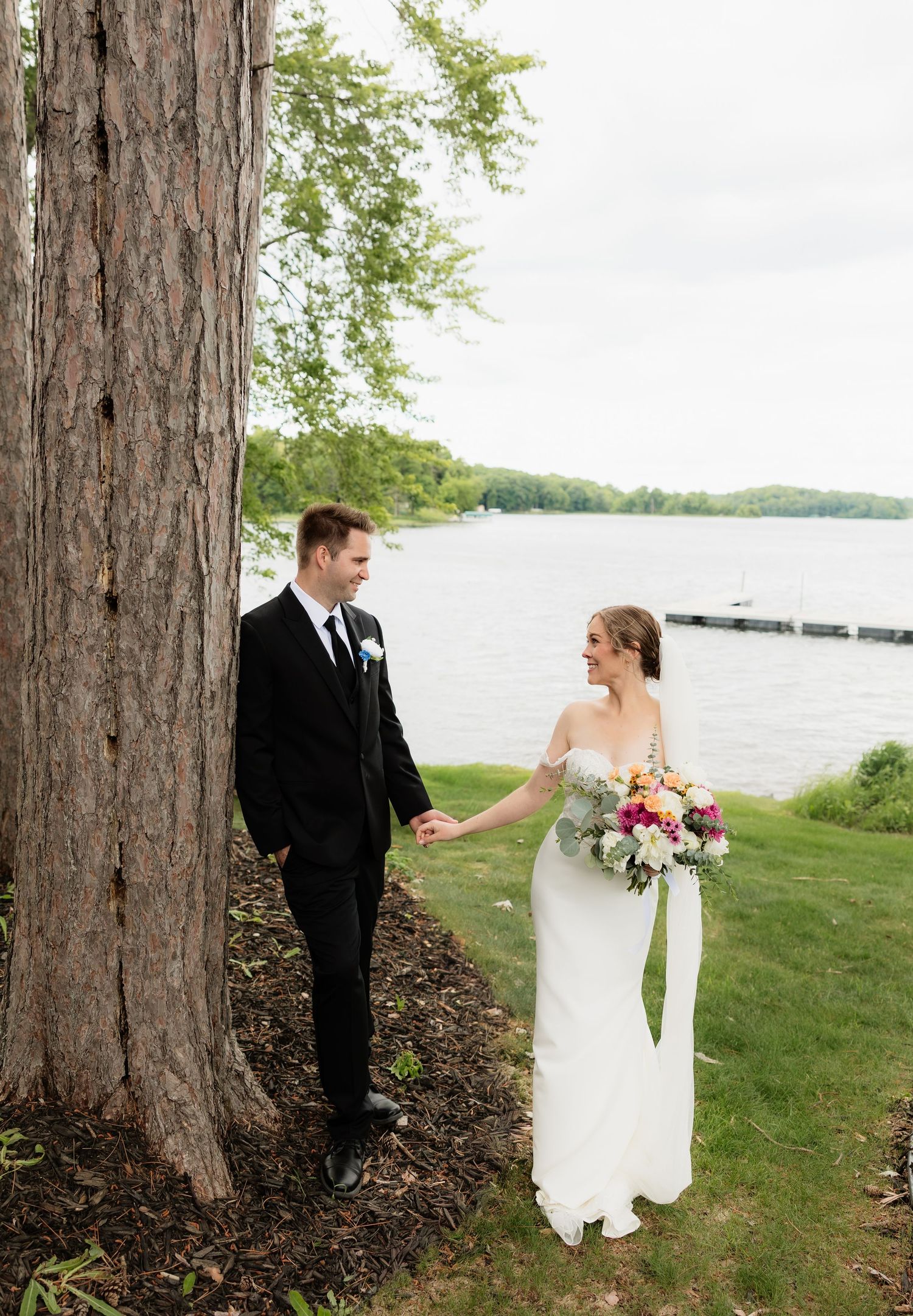Bride and groom holding hands by a large tree near a lakeside dock, bride holding a colorful bouquet in a white gown.