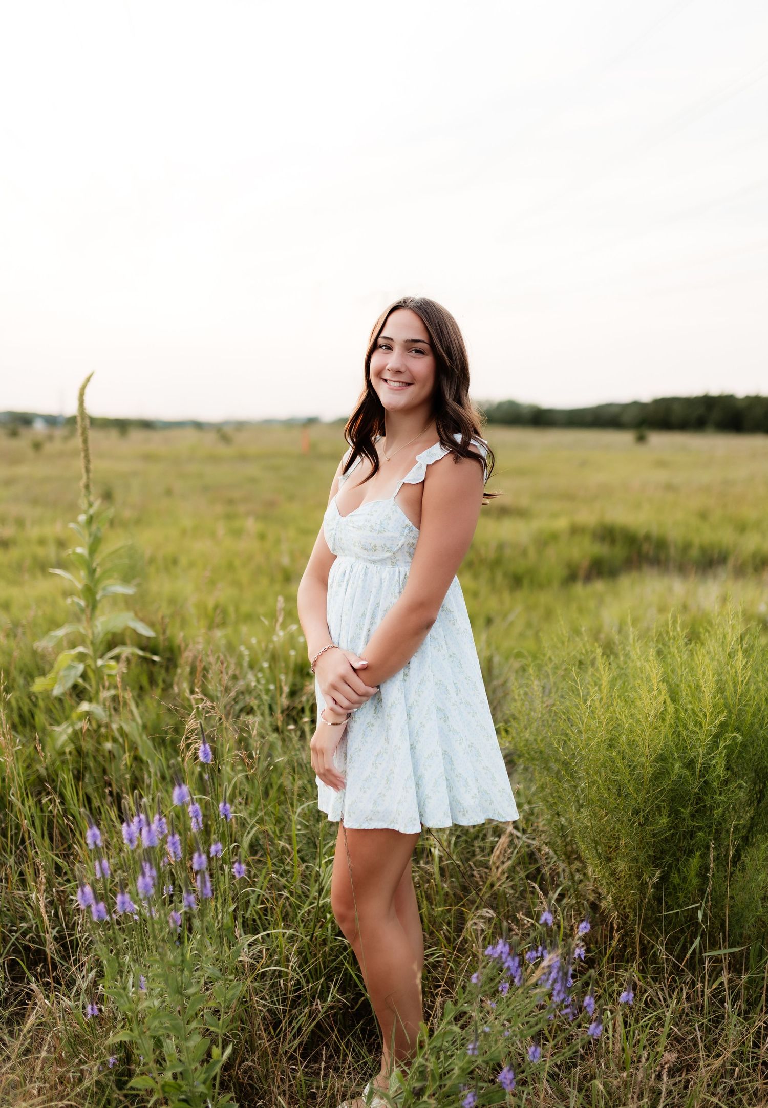 Smiling young woman in light blue mini dress standing in wildflower meadow at golden hour.