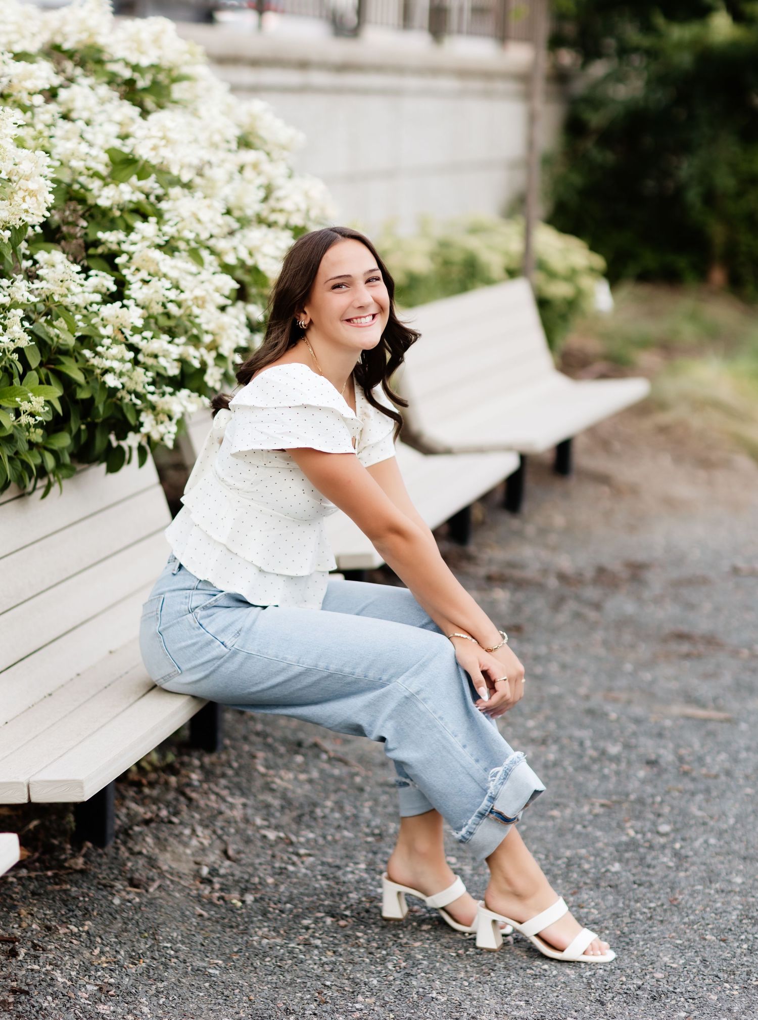 Young woman in white ruffled top and jeans sits smiling on a white bench beside blooming white flowers.