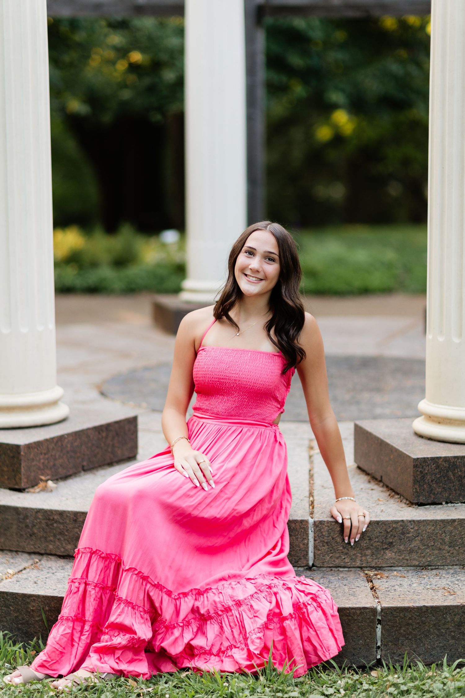 Young woman in pink maxi dress sitting on stone steps between white columns.