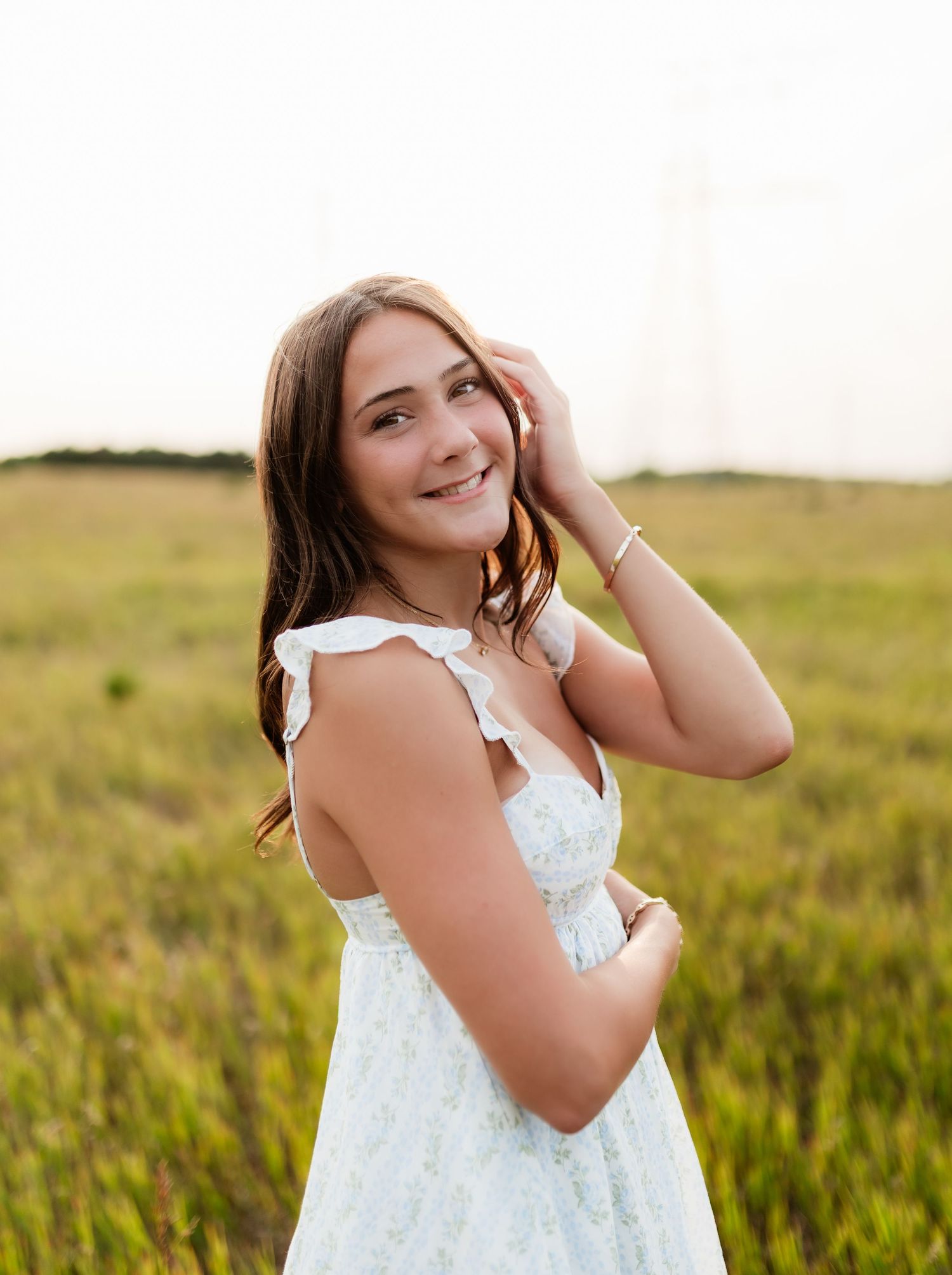 Young woman in white floral dress smiling in golden grass field at sunset.