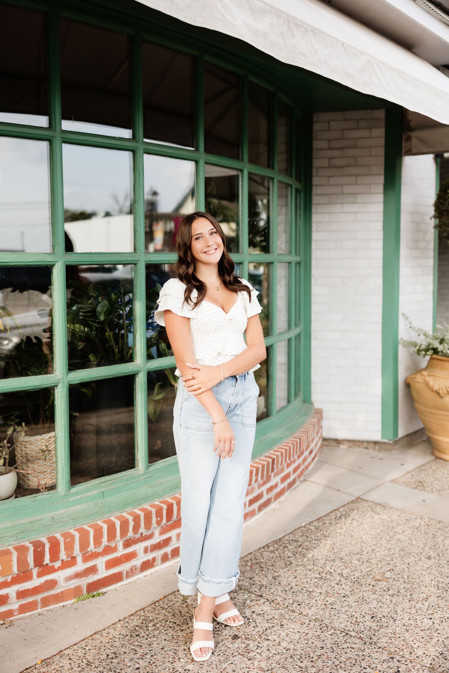 Young woman in white top and light jeans smiling outside a brick building with green windows.