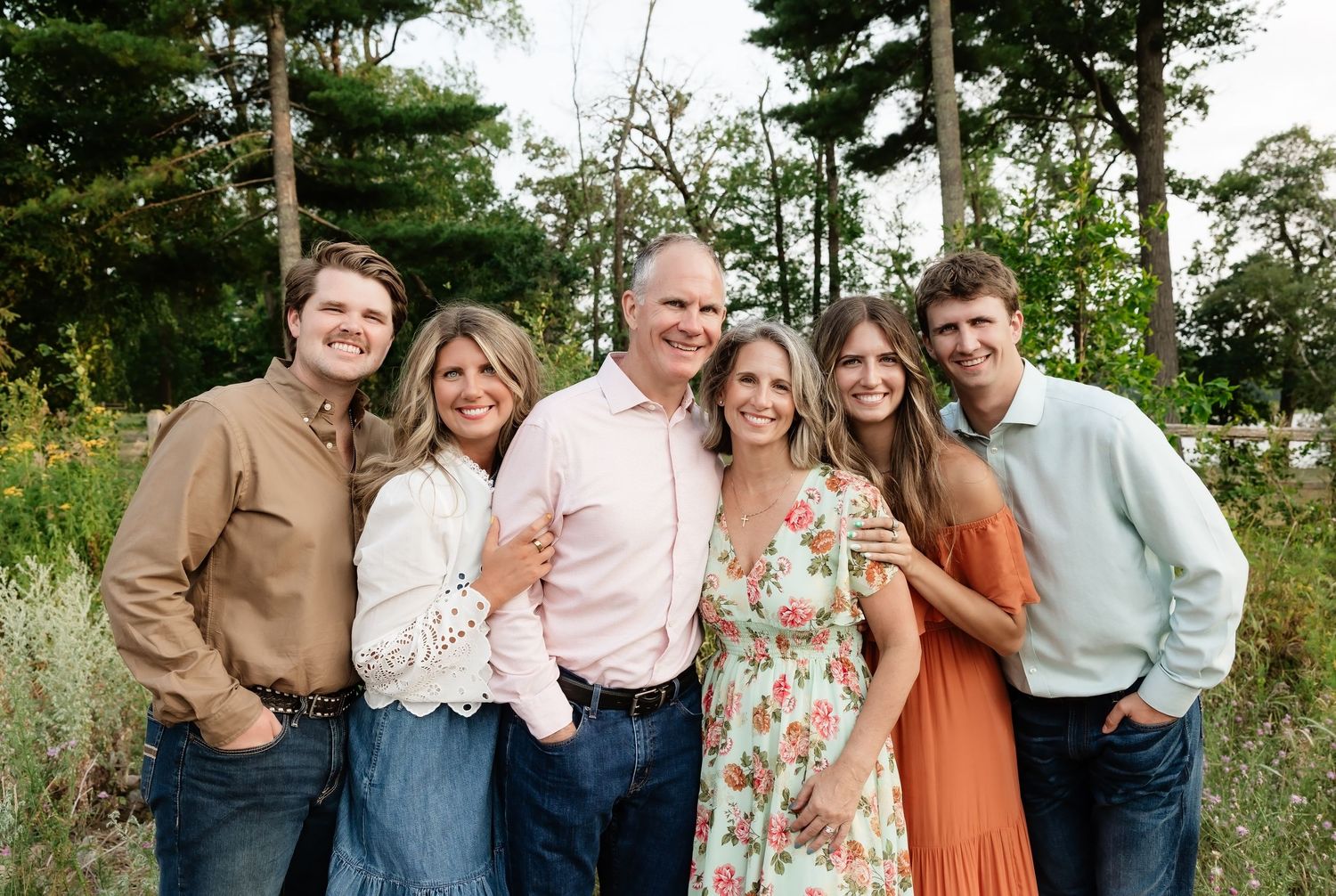 Six family members smile together outdoors in a lush green setting, dressed in coordinated casual outfits.