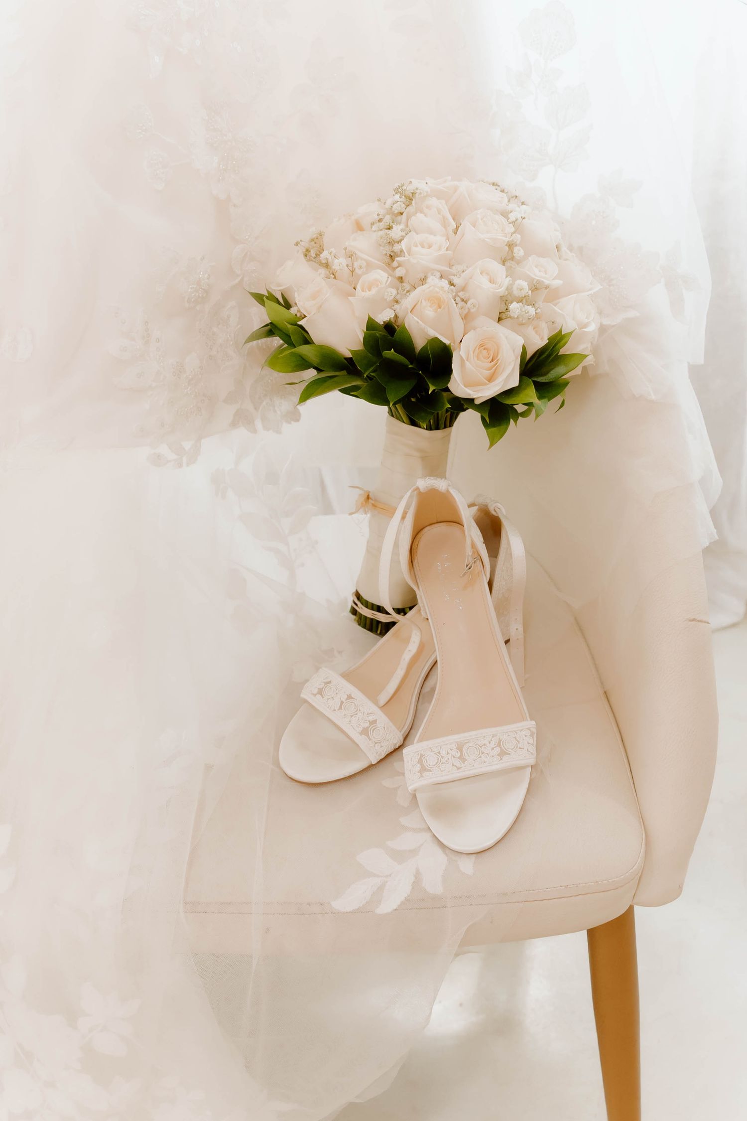 White bridal heels and rose bouquet displayed on a chair beside a lace wedding gown.