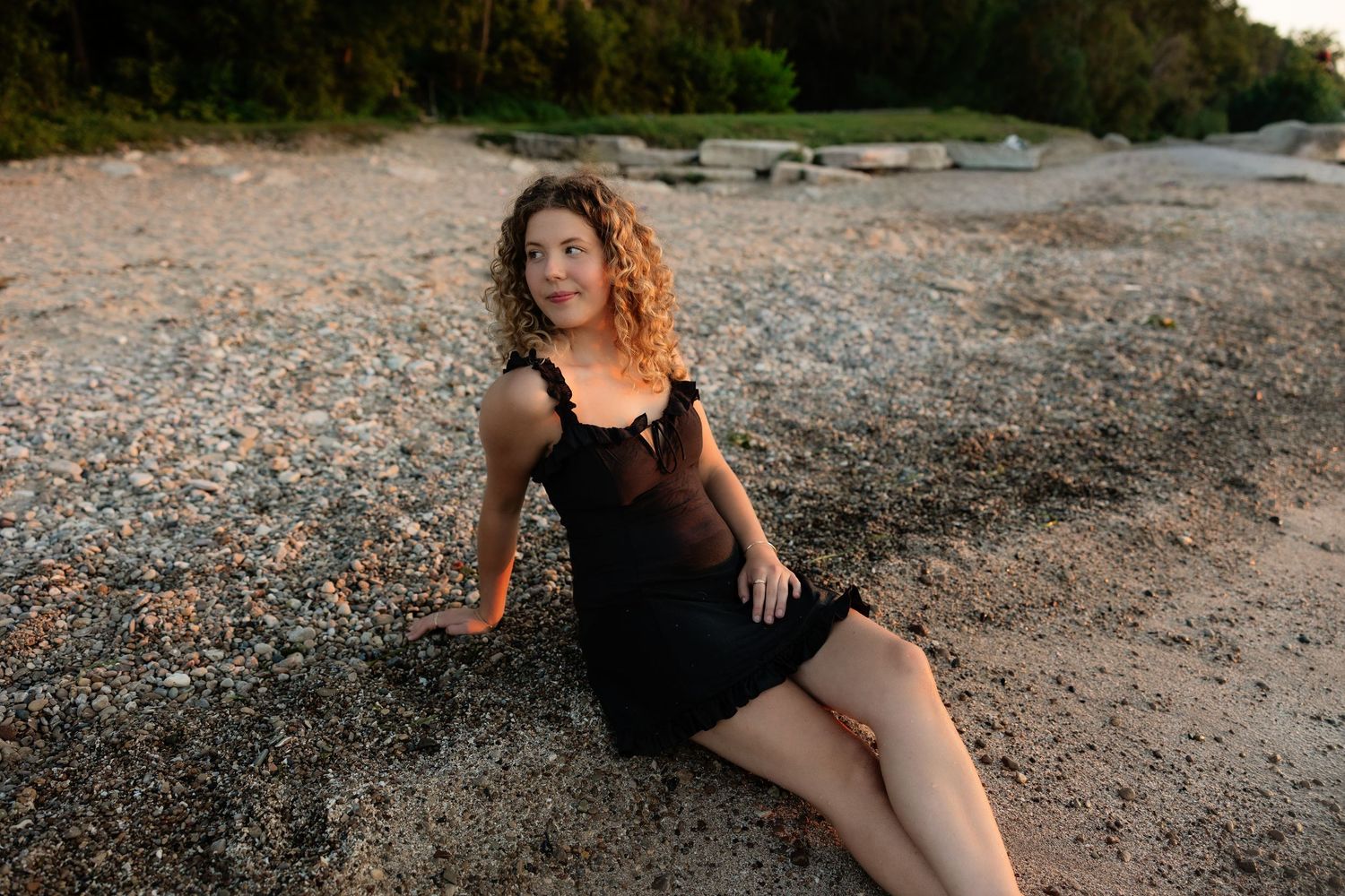 Young woman with curly hair in black mini dress posing on pebble beach at dusk.