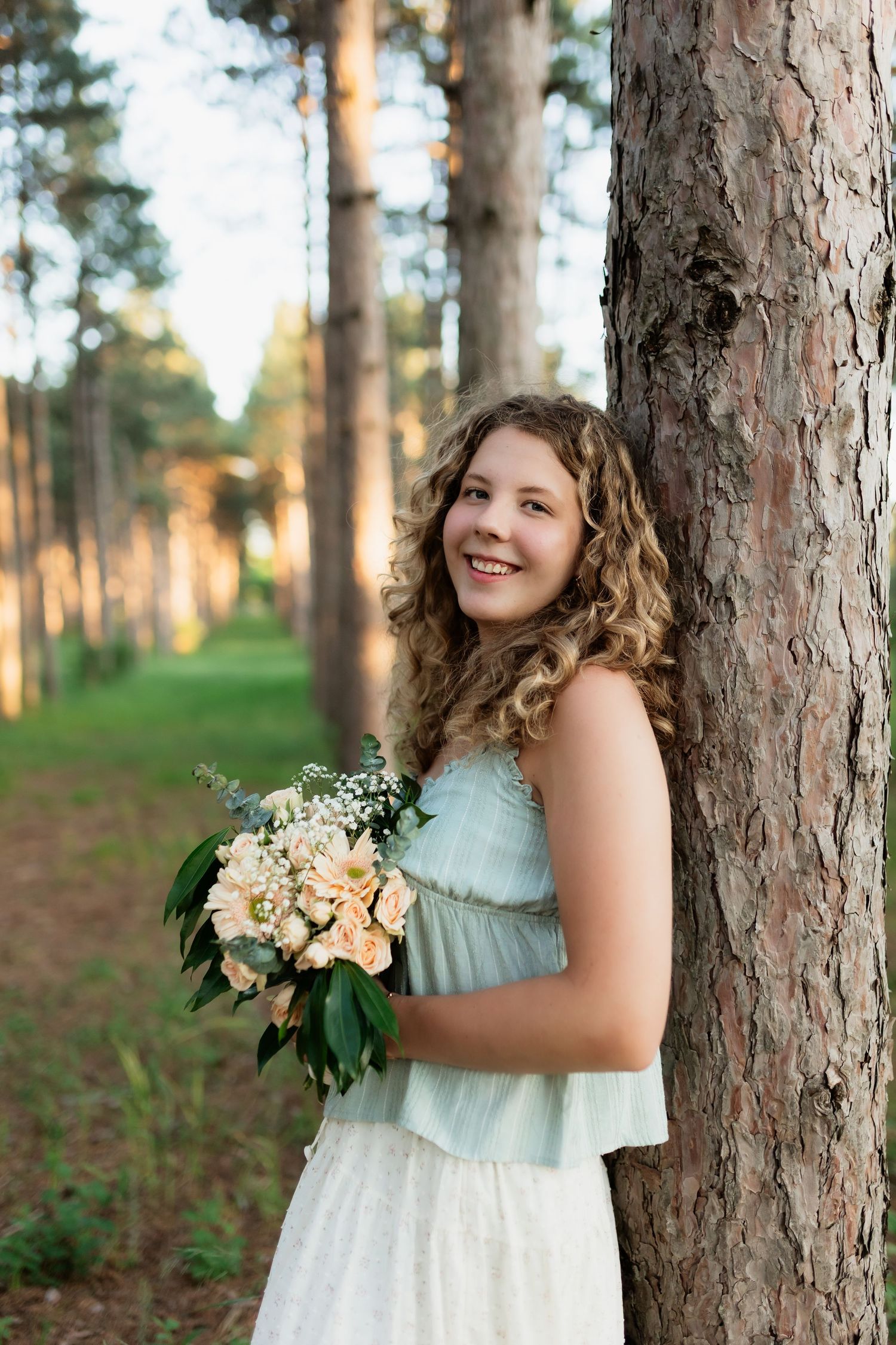 Smiling curly haired girl leaning against pine tree holding flower bouquet.