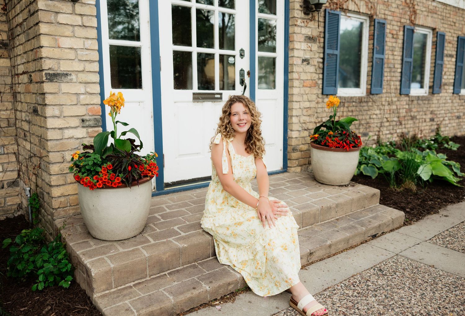 Young woman in floral dress sitting on brick steps with colorful potted flowers outside a charming home.