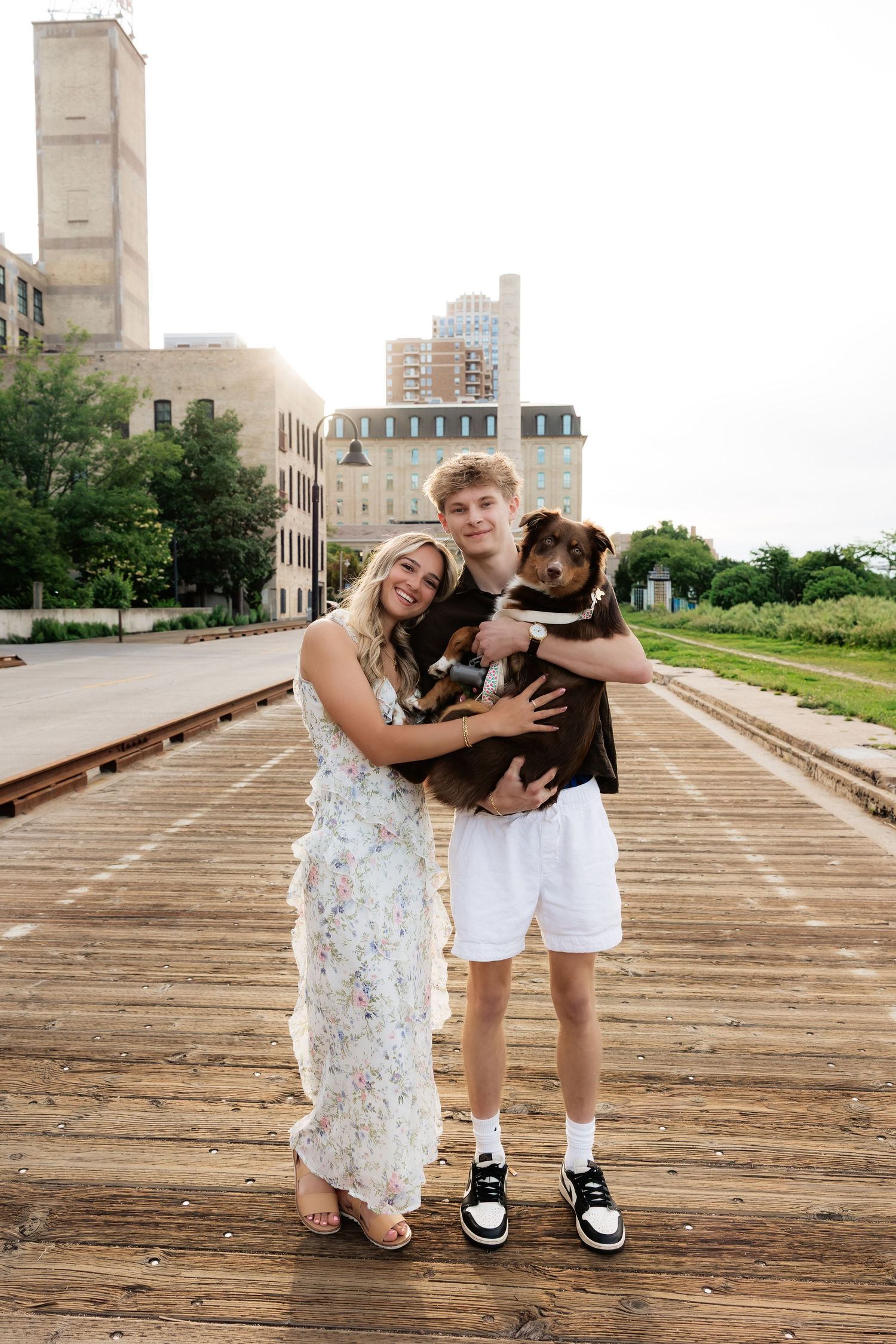 Young couple smiling on wooden railroad bridge holding brown dog with urban buildings in background.