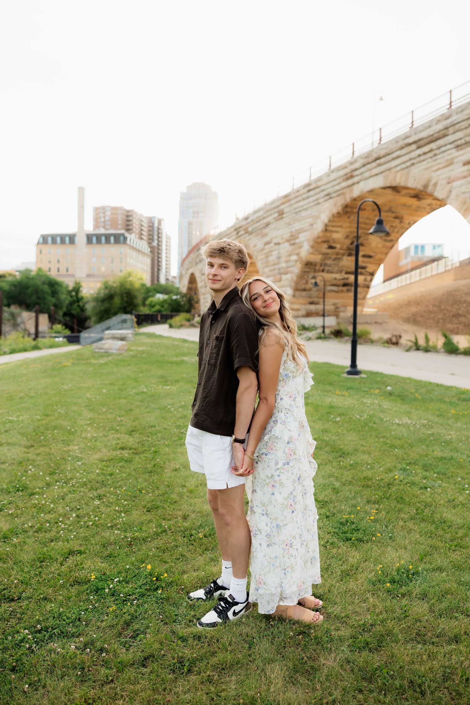 Couple posing back-to-back on green grass with a scenic stone arch bridge in the background.