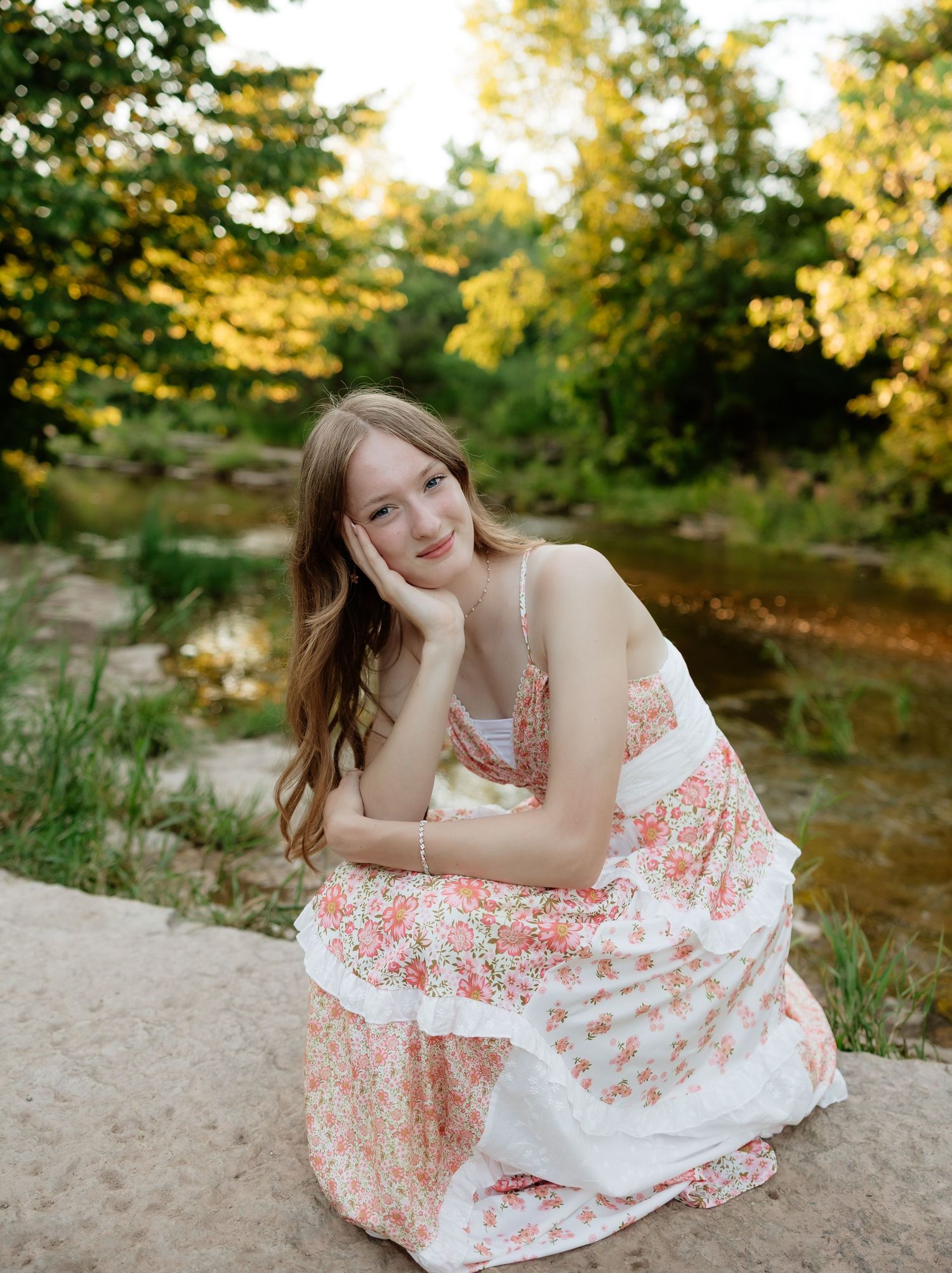Young woman in a floral dress posing by a creek in a golden hour outdoor portrait.