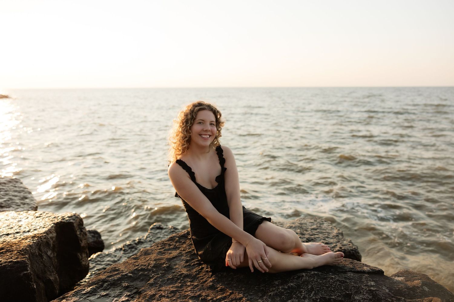 Woman with curly hair in black dress sitting on rocks by the ocean at sunset, smiling.