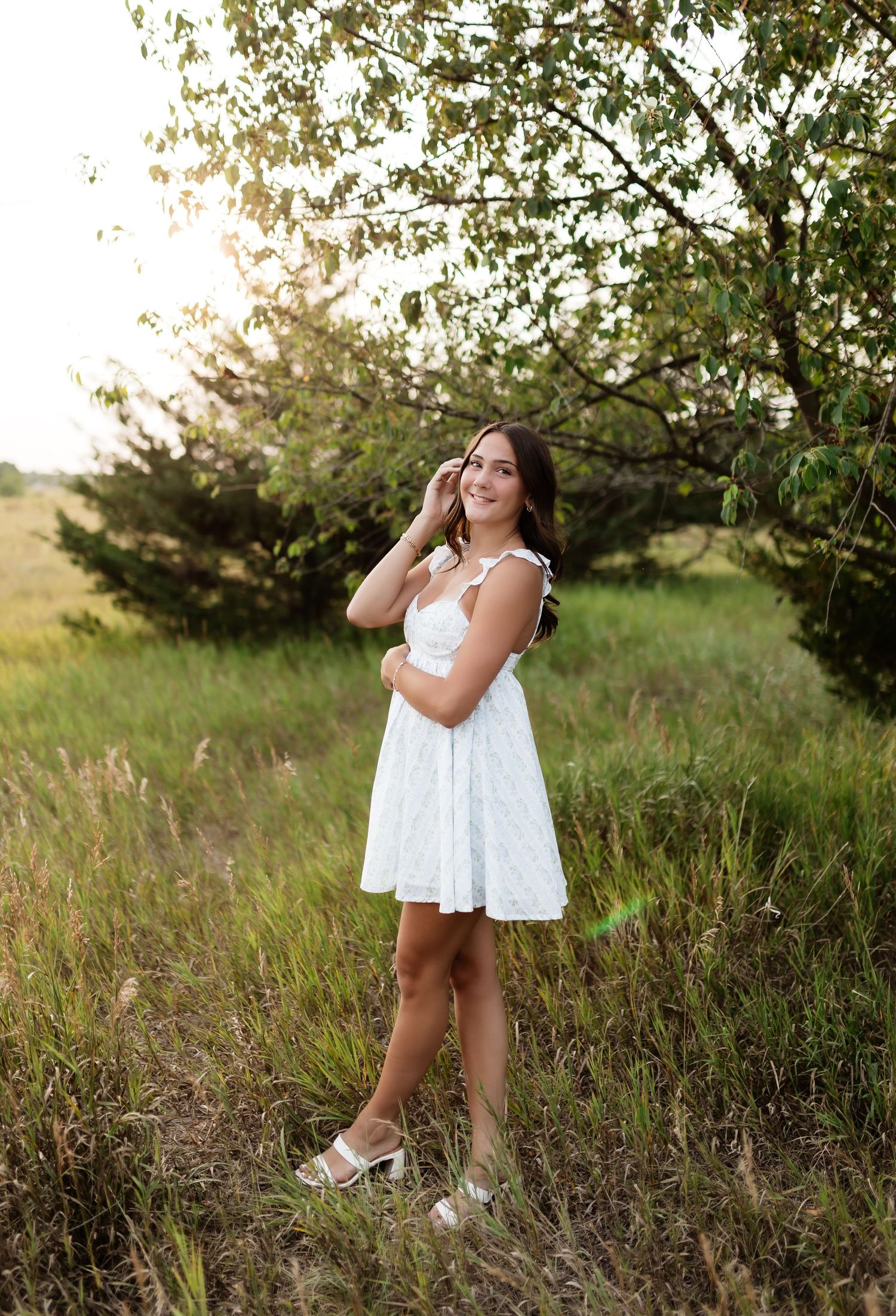 Young woman in white mini dress standing in sunlit meadow near trees.