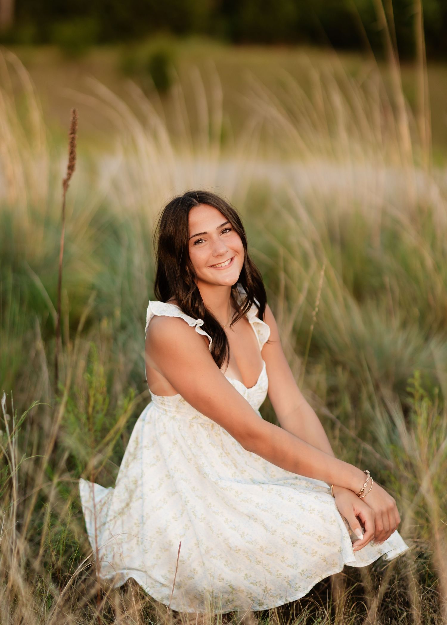 Smiling woman in white floral dress sitting in tall golden grass field.