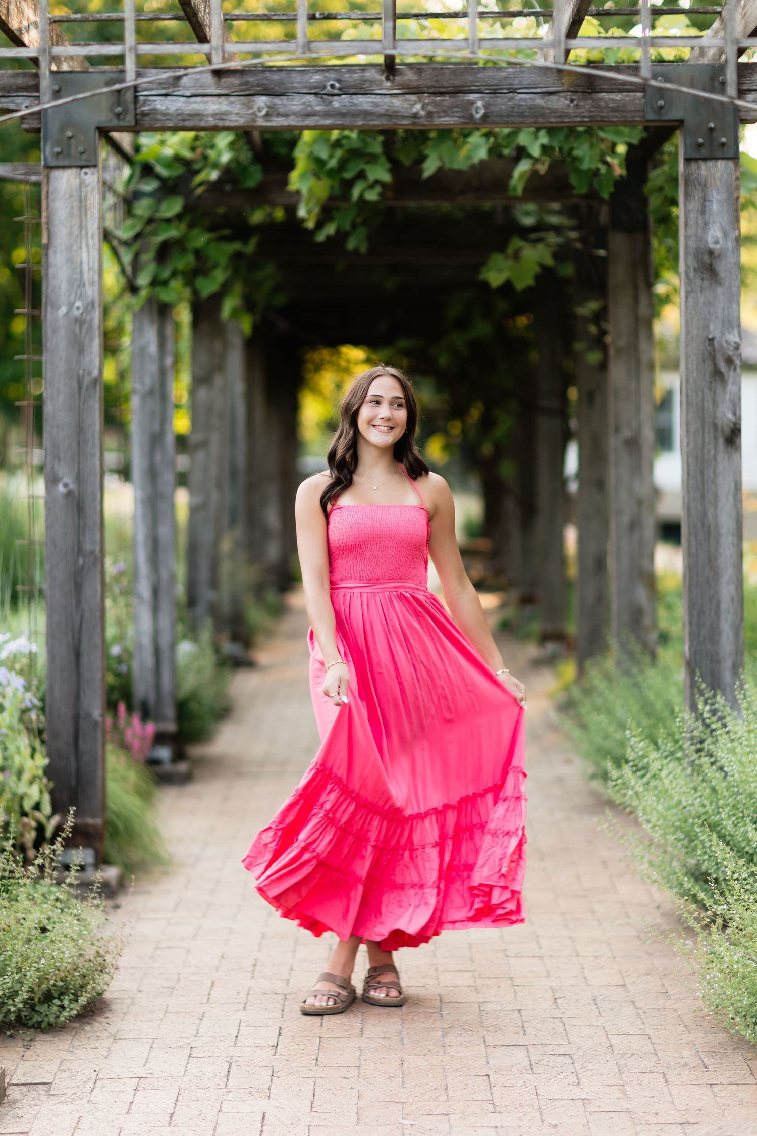 Smiling woman in pink dress walking under a wooden pergola in a garden path.