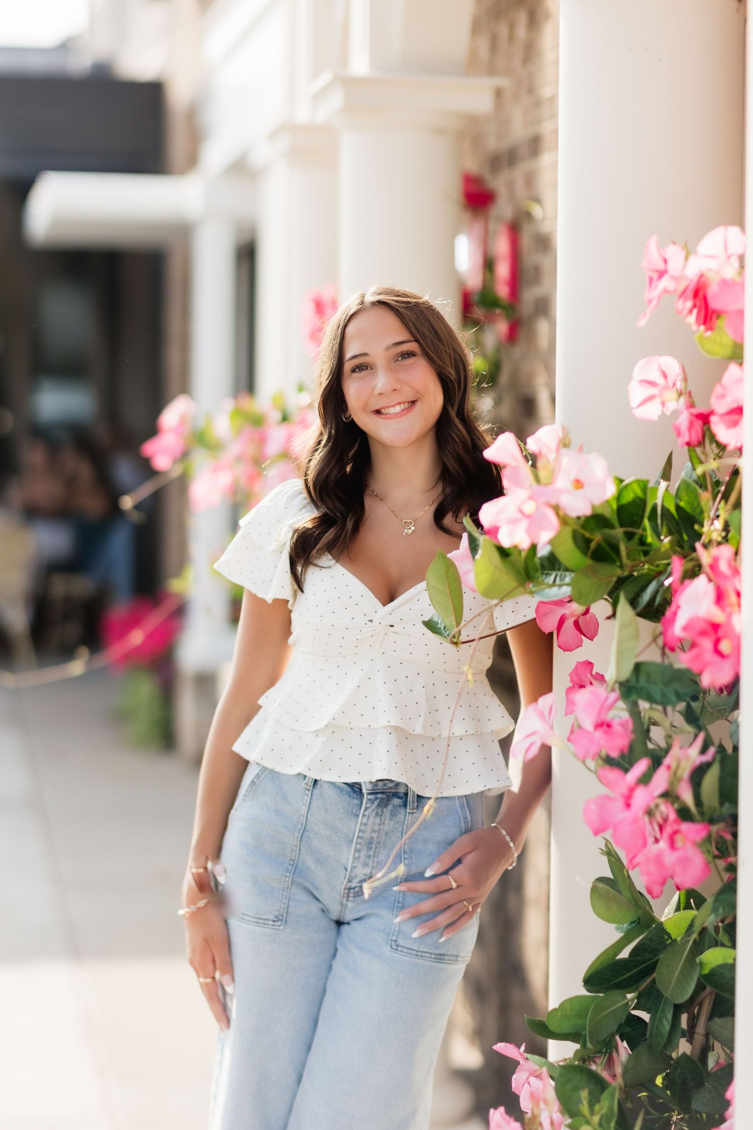 Smiling brunette woman in white eyelet top and blue jeans standing beside vibrant pink flowers on a sunny day.