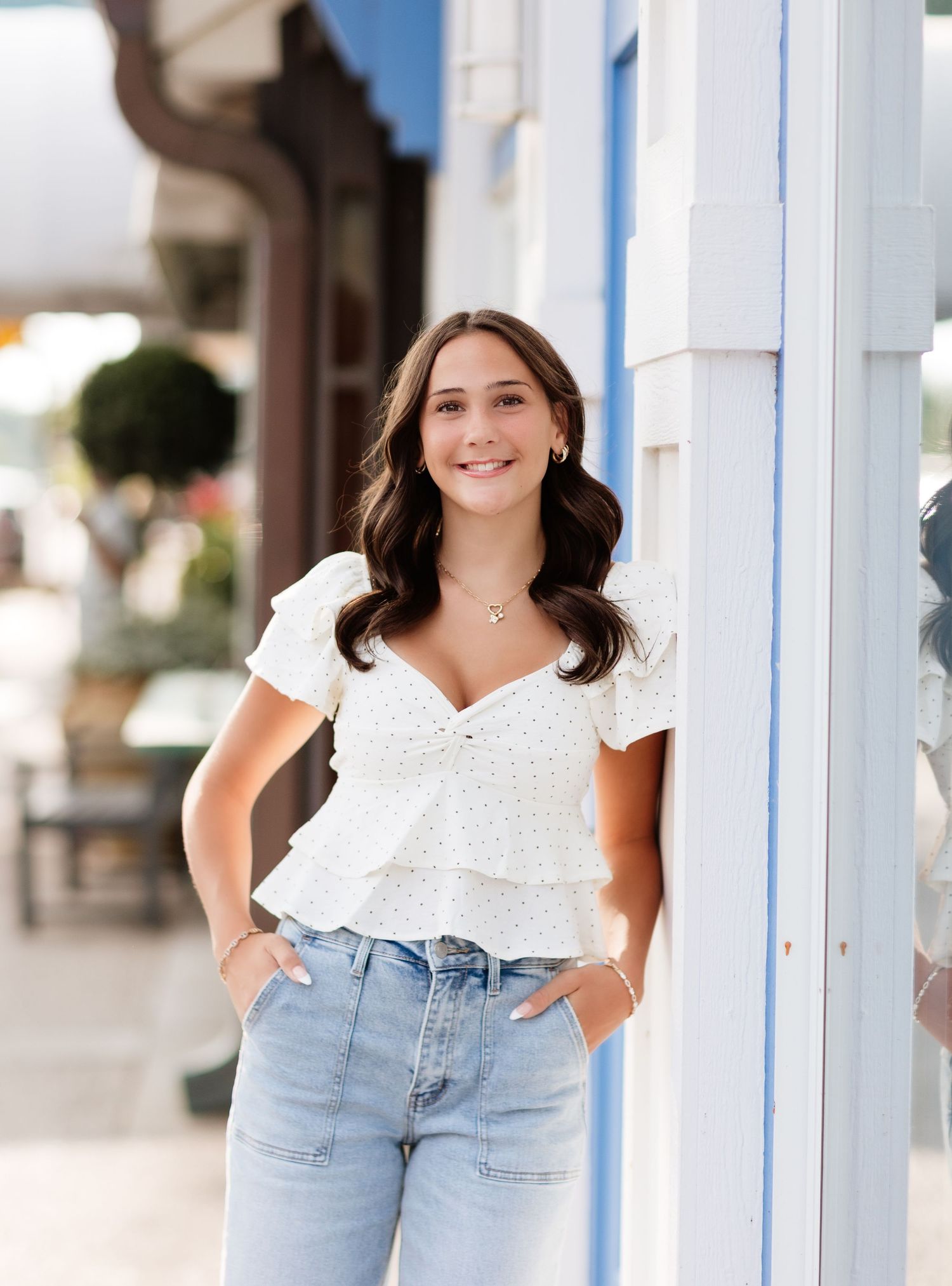 Young woman in white ruffled top and jeans smiling outdoors near a blue and white storefront.