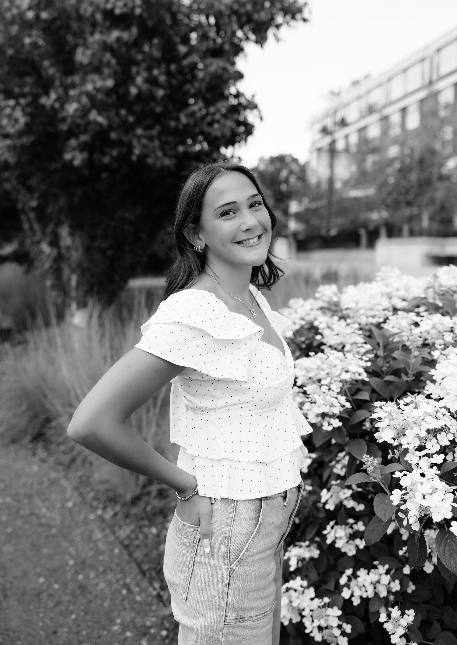 Young woman smiling in black and white photo, wearing ruffled white top, standing beside blooming flowers in a garden.