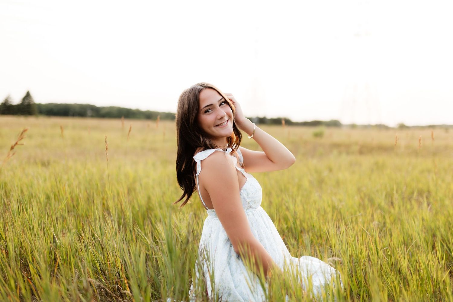 Young woman in white dress sitting in a golden grass field, laughing and touching her hair.