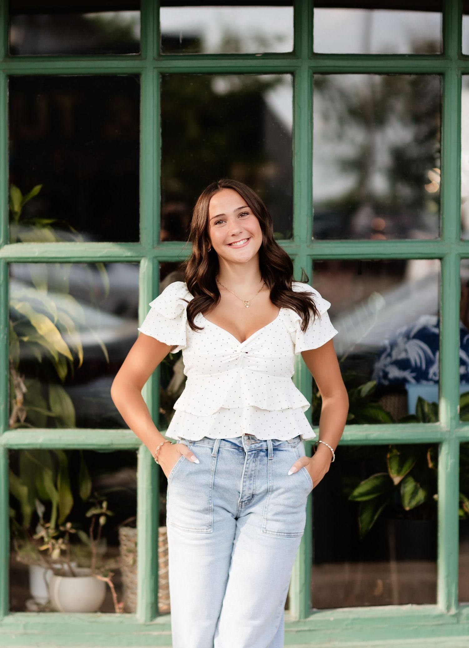 Smiling teen girl in white peplum top and jeans standing before green-framed window with plants.