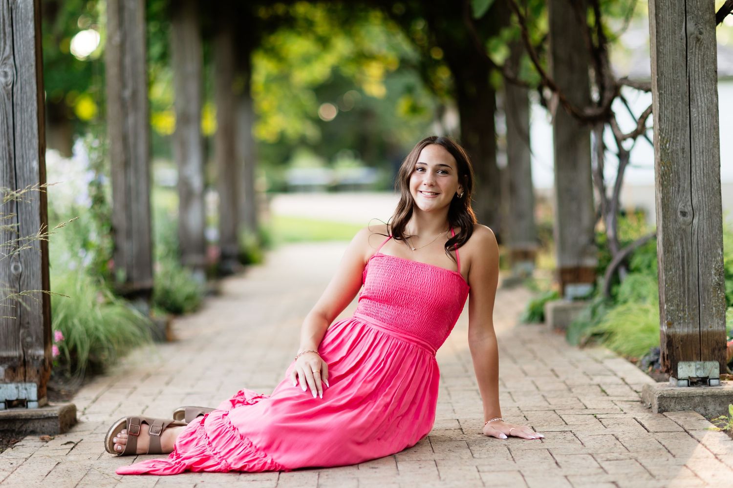 Young woman in pink maxi dress sitting on brick path under vine-covered pergola.