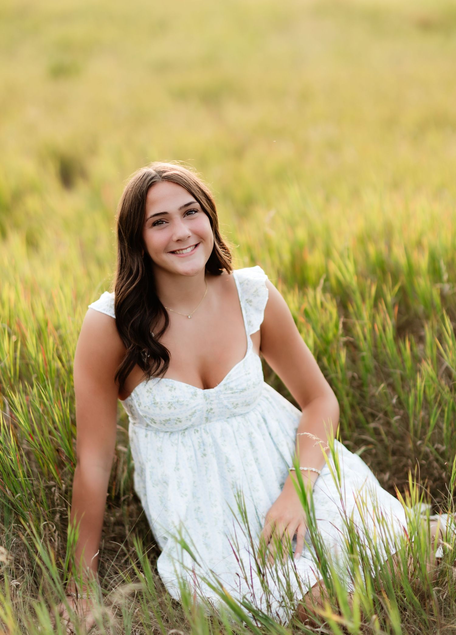 Young woman in white floral dress sitting in golden grass field, smiling outdoors.