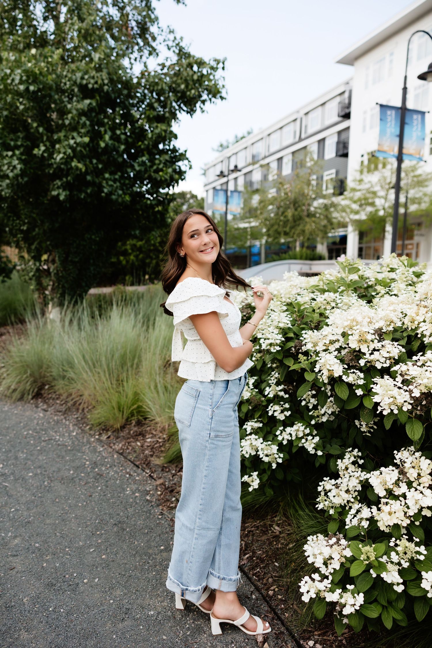 Young woman in white top and jeans smiling beside white flowering bushes in an urban garden setting.
