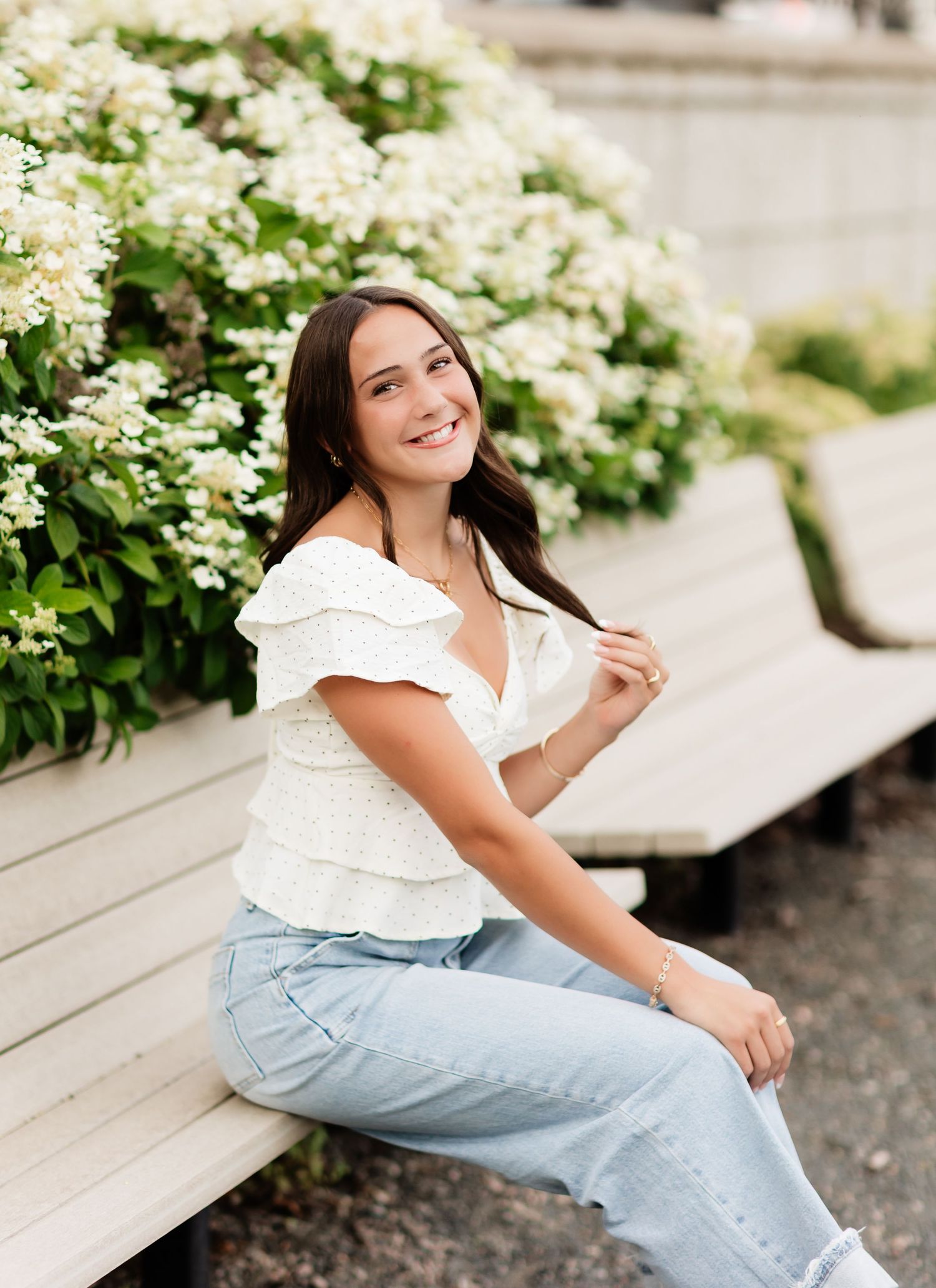 Young woman with dark hair smiles while sitting on a bench surrounded by white flowering bushes.