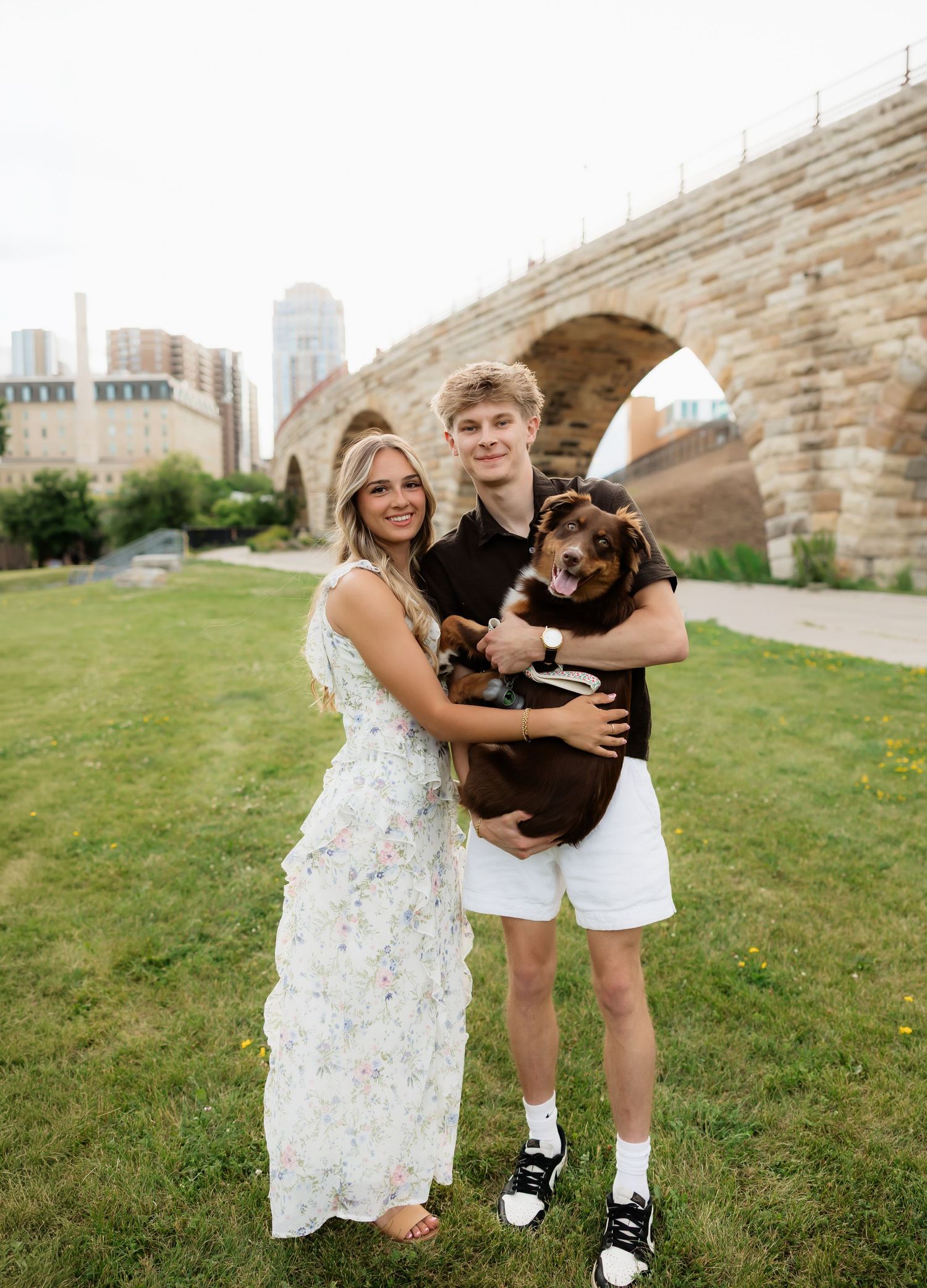 Smiling couple holding brown dog on green lawn with stone arch bridge and city skyline backdrop.