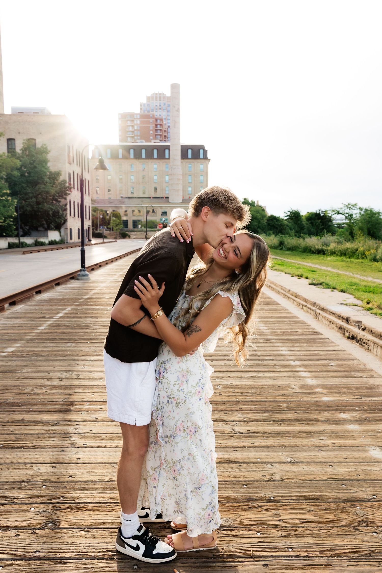 Man kissing woman on the cheek as she laughs on a wooden boardwalk with city buildings behind.