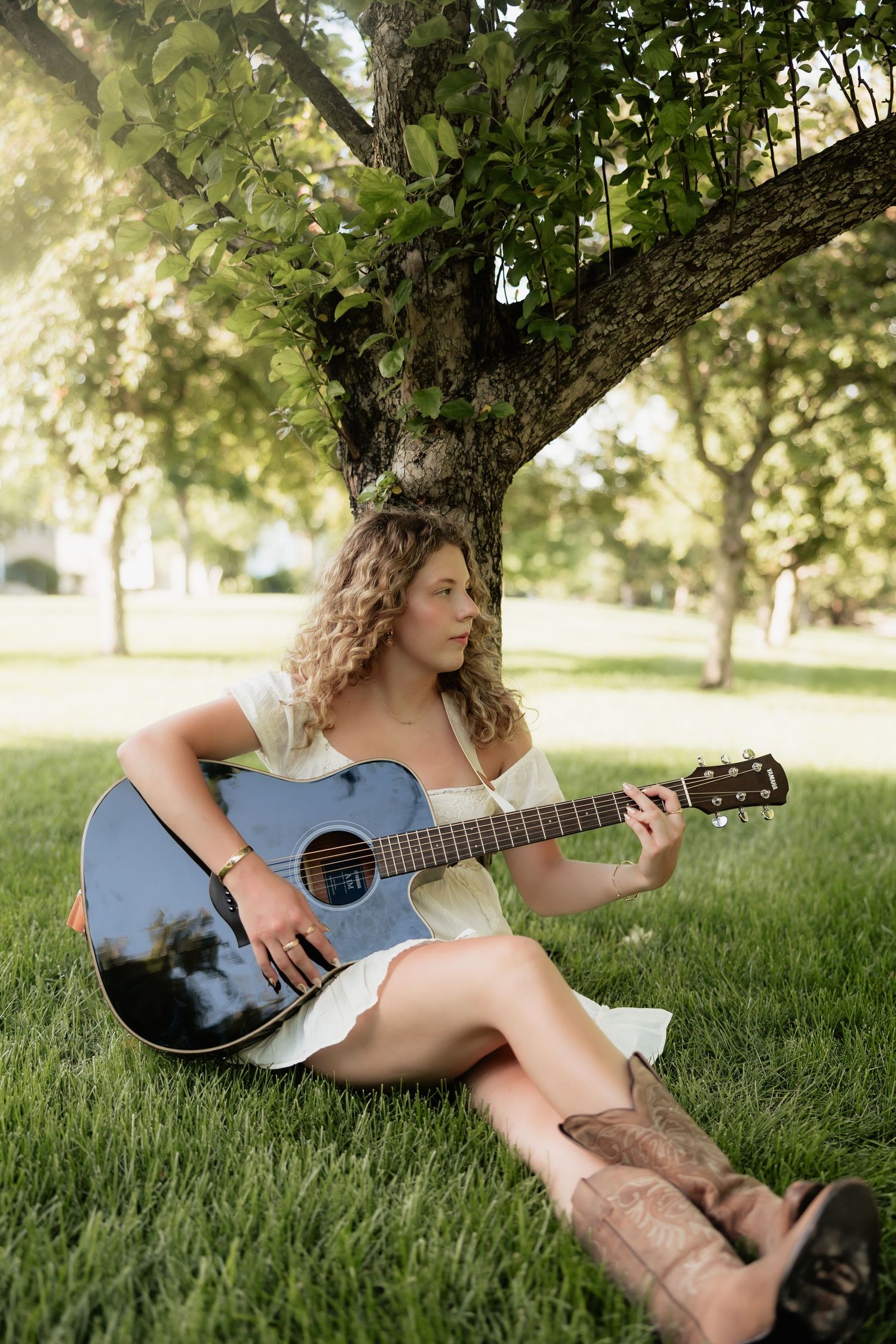 Young woman with curly hair playing acoustic guitar while sitting on grass under a tree in sunlight.