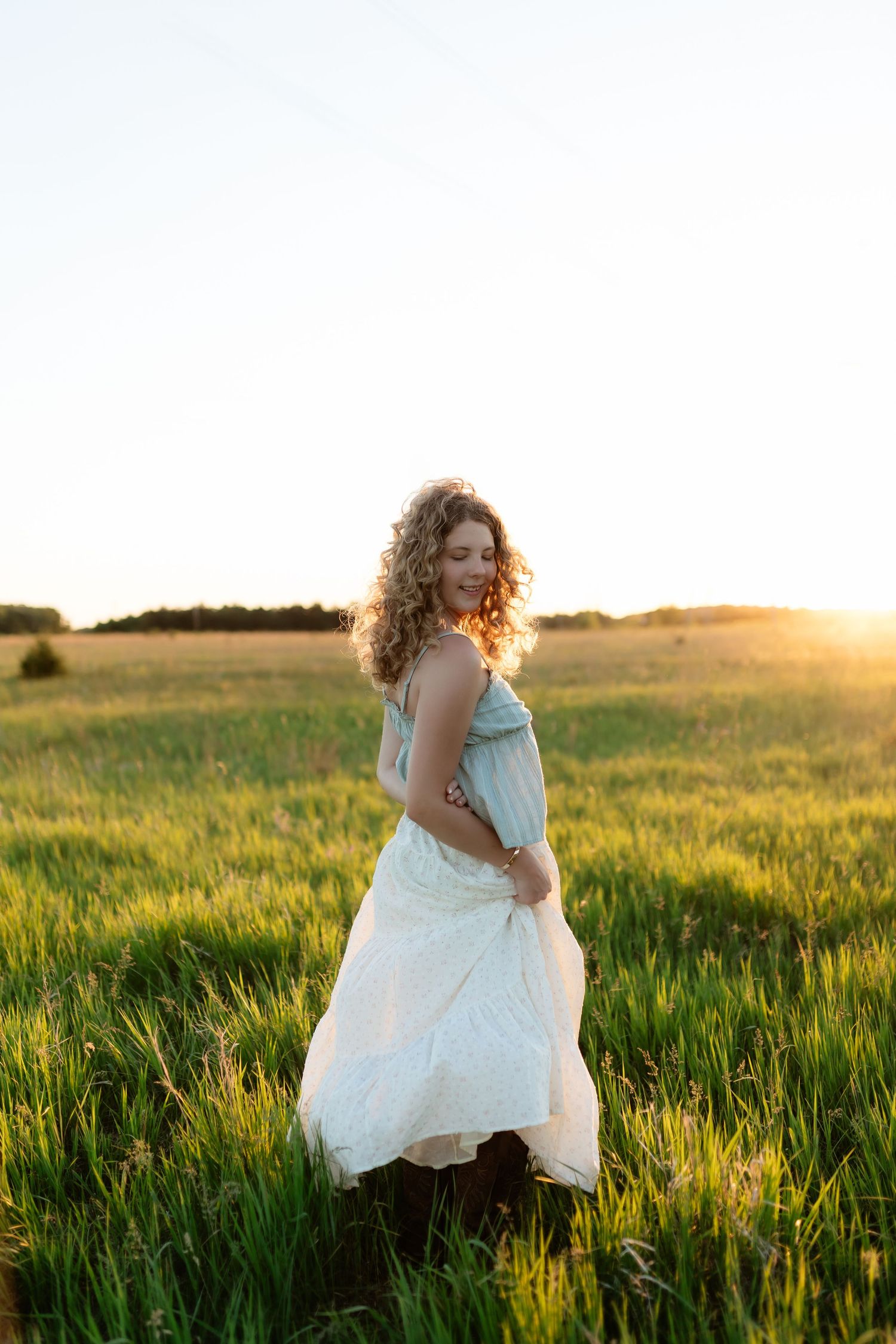 Curly-haired woman in flowing white dress standing in green field at golden sunset.