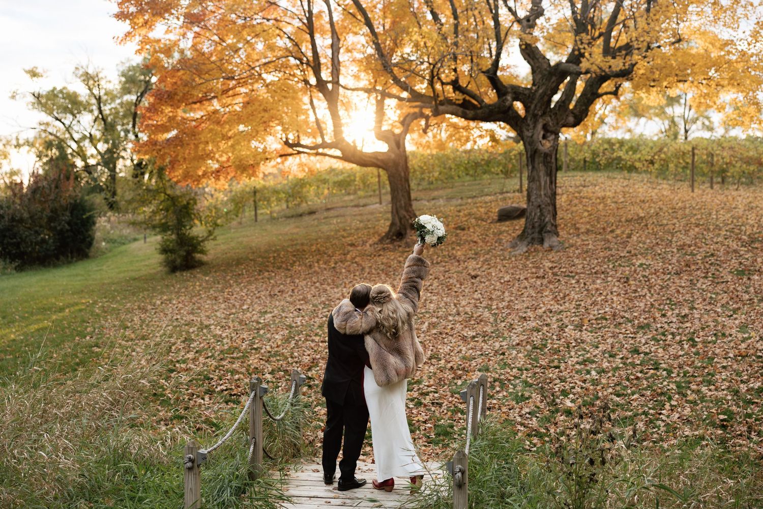 Bride and groom walking hand in hand on a wooden bridge surrounded by golden autumn leaves at sunset.