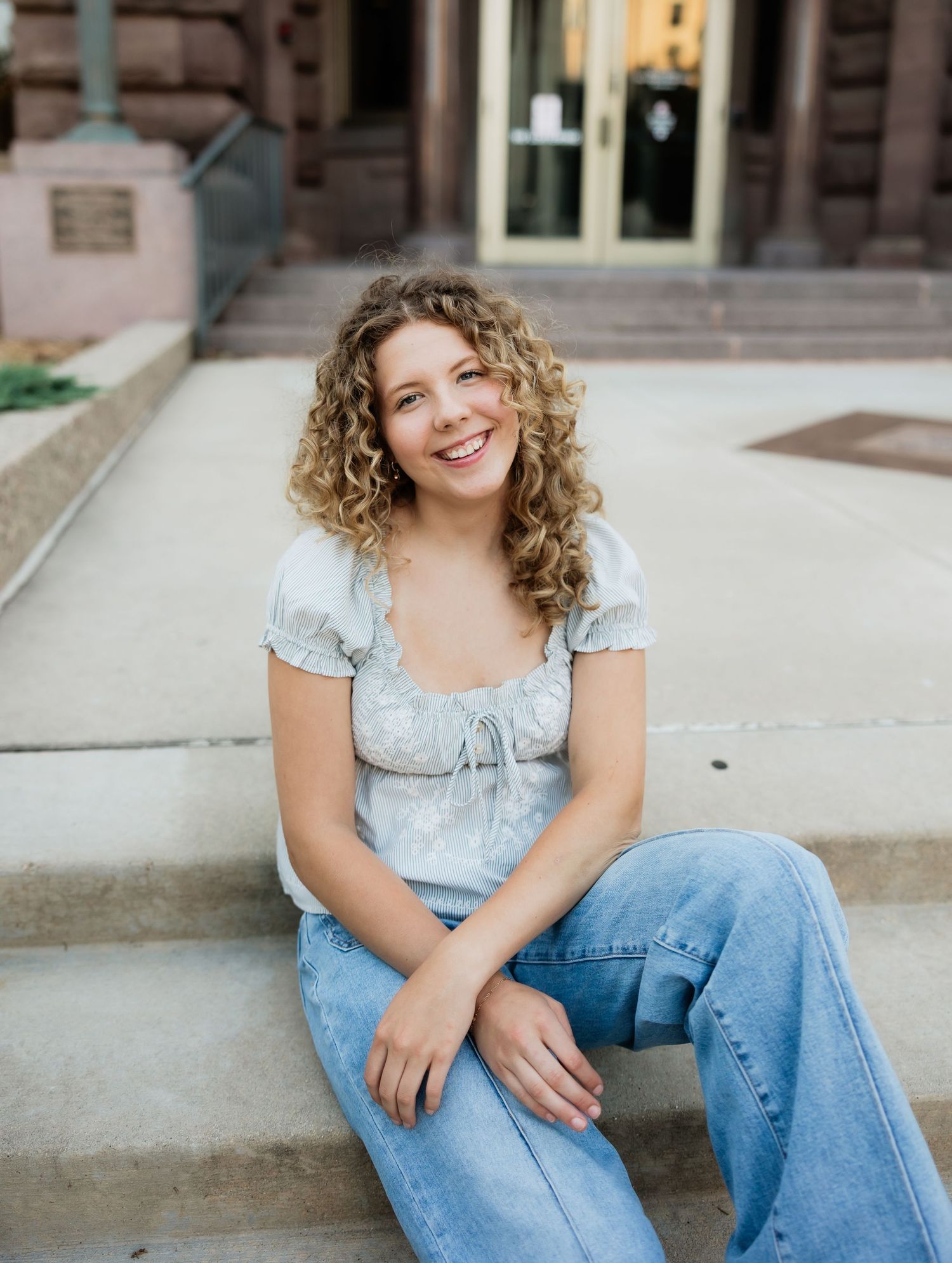 Young woman with curly hair smiling on city steps wearing a floral top and jeans.