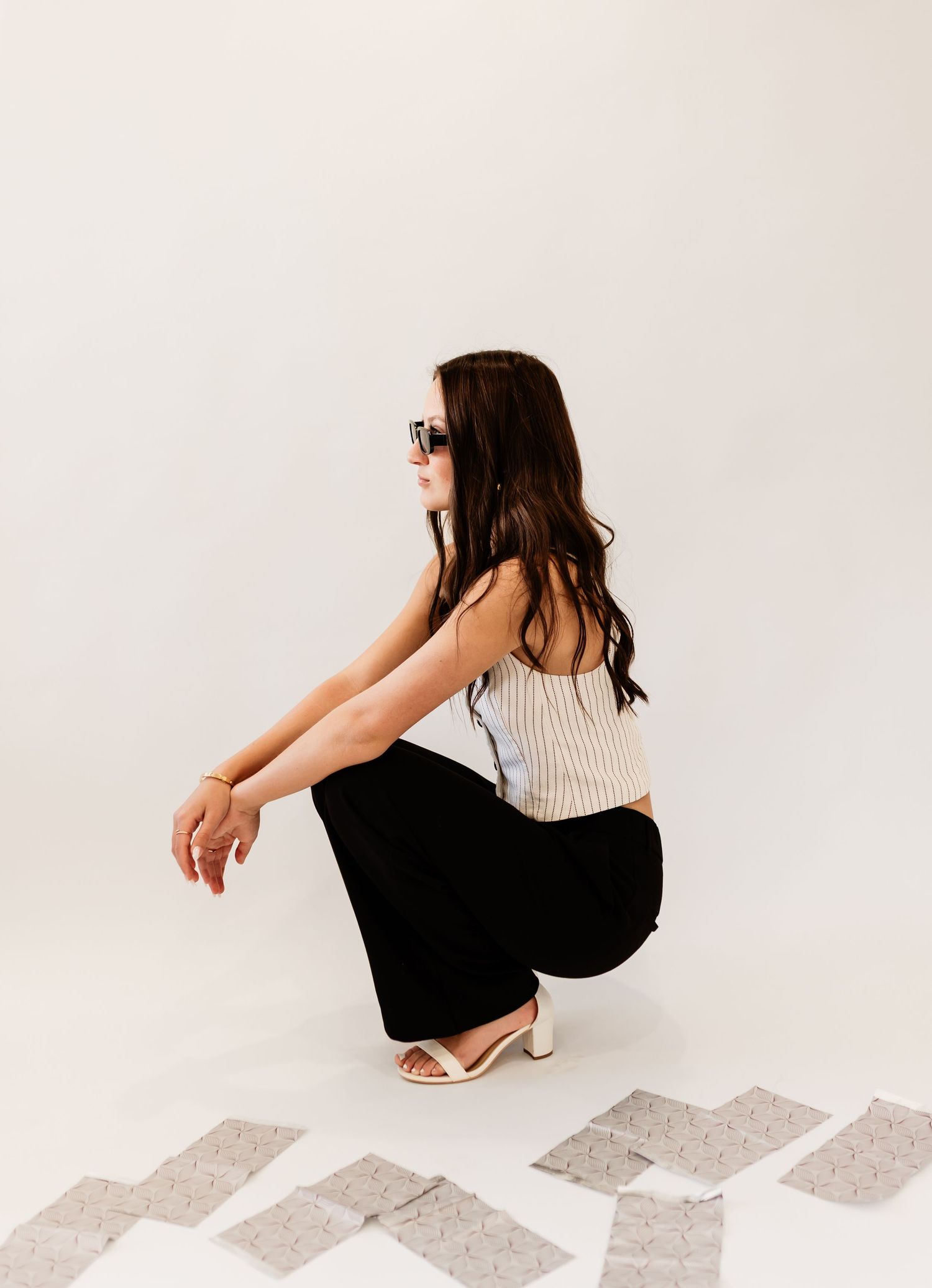 Brunette woman in sunglasses, striped crop top, and black wide-leg pants crouching in white studio.