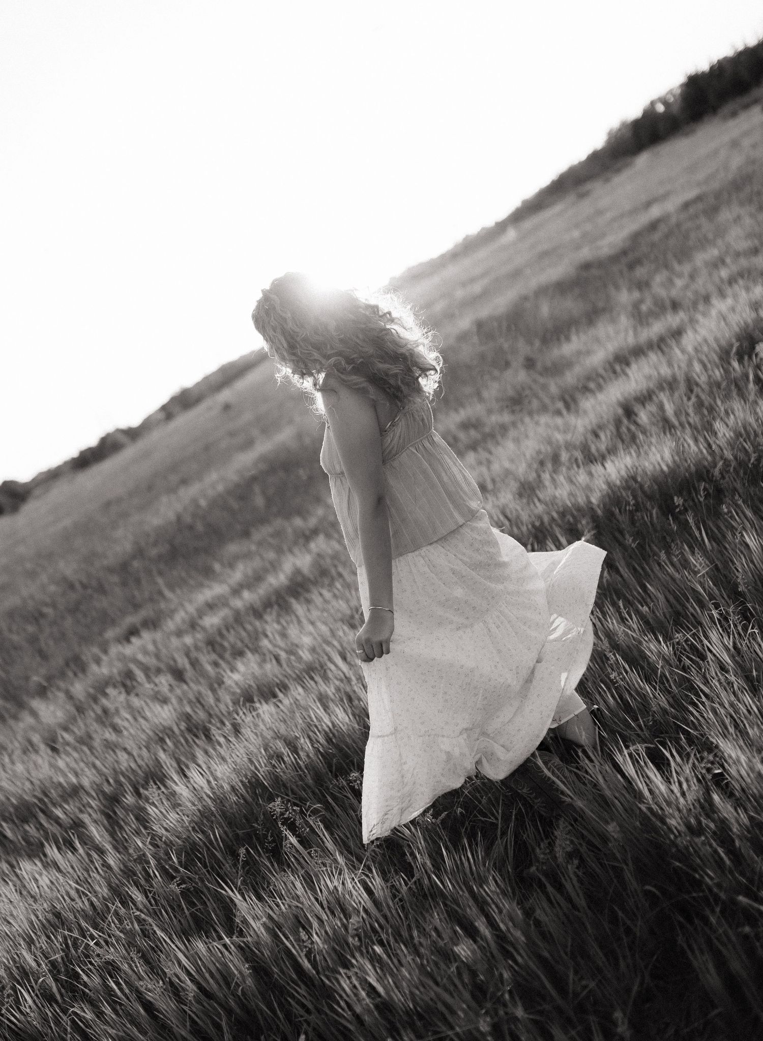 Black and white photo of a woman in a flowing dress walking through a sunlit grassy hillside field.