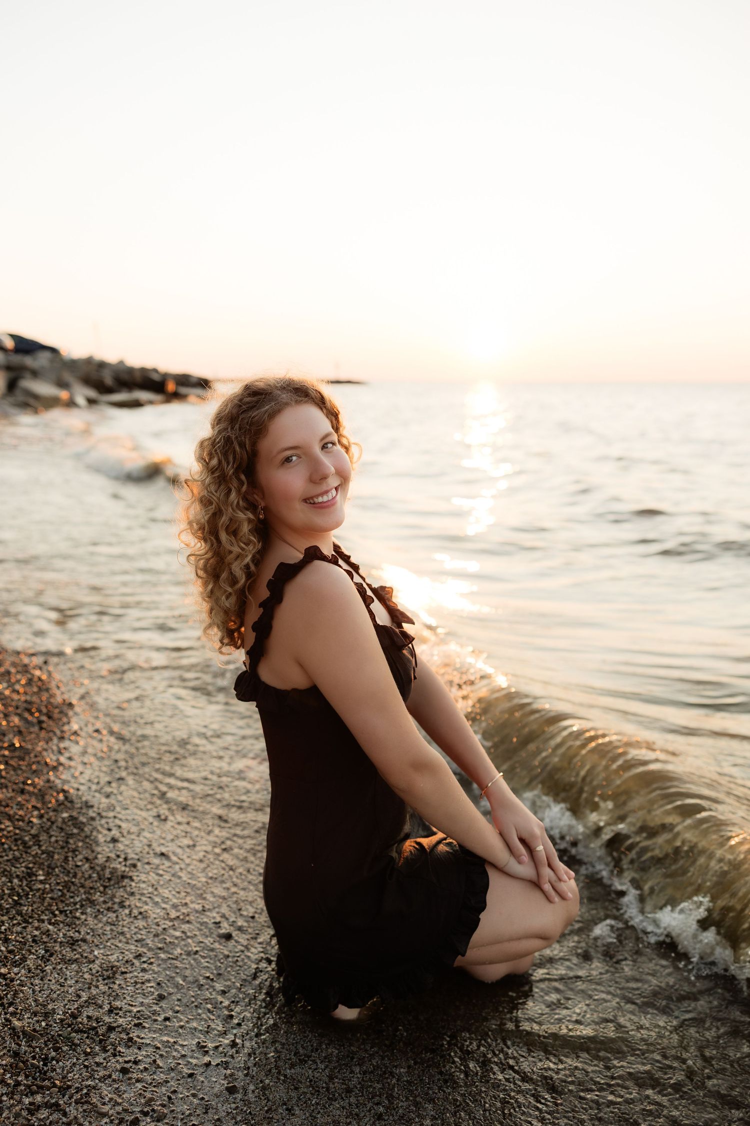 Young woman with curly hair in black outfit kneeling on beach shore smiling at sunset.
